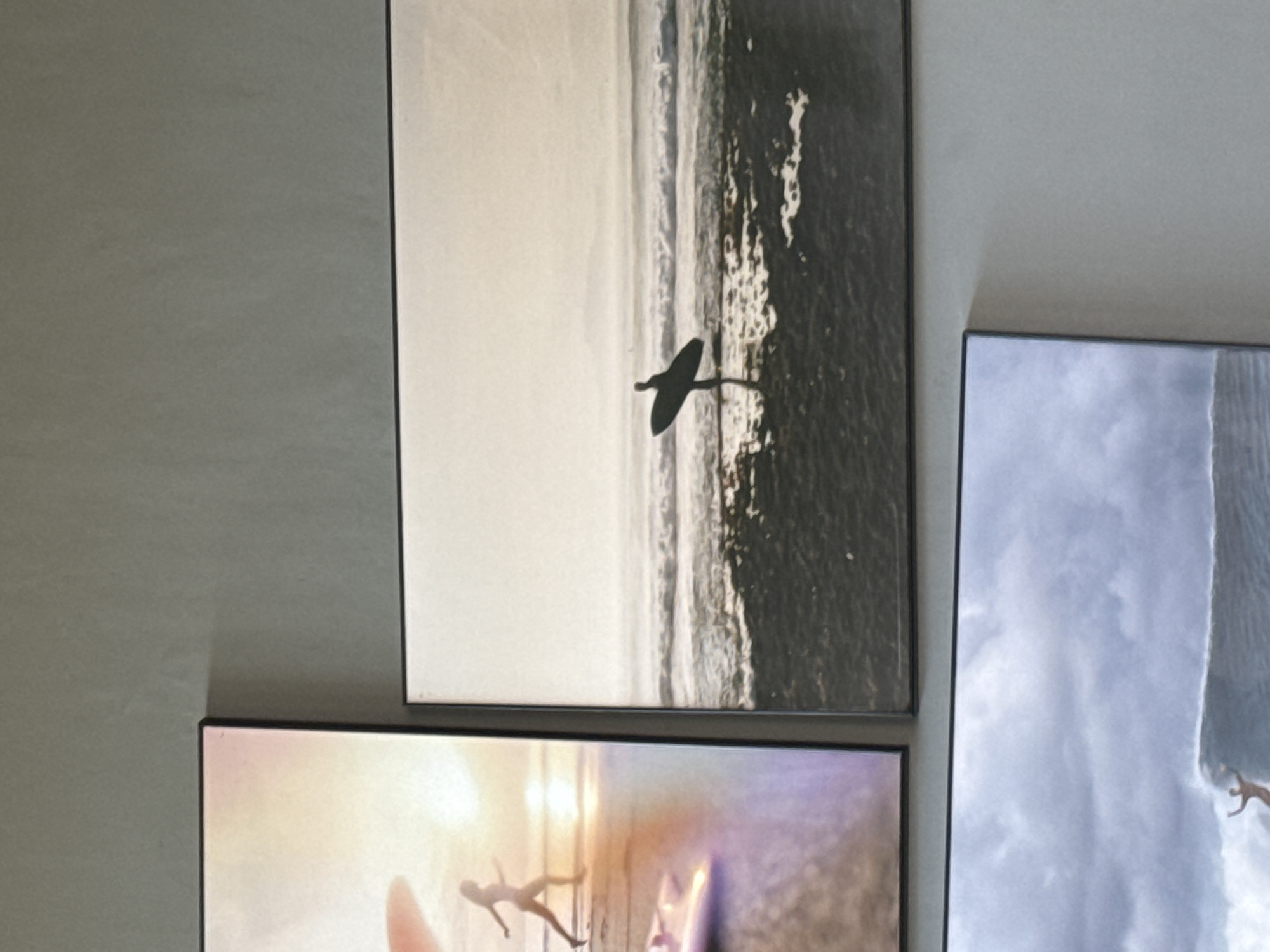 The image shows a section of a wall adorned with framed artworks. The top right picture is a monochrome scene of a coastline, showing a beach and sea with a bird flying over the waves. Below this is a photograph that features what appears to be a beach scene with human figures. Another picture is visible to the right, with a cloudy-sky scene.
Given the "Kuta Selatan, Indonesia" prompt, the beach scenes possibly represent beaches in that location.
