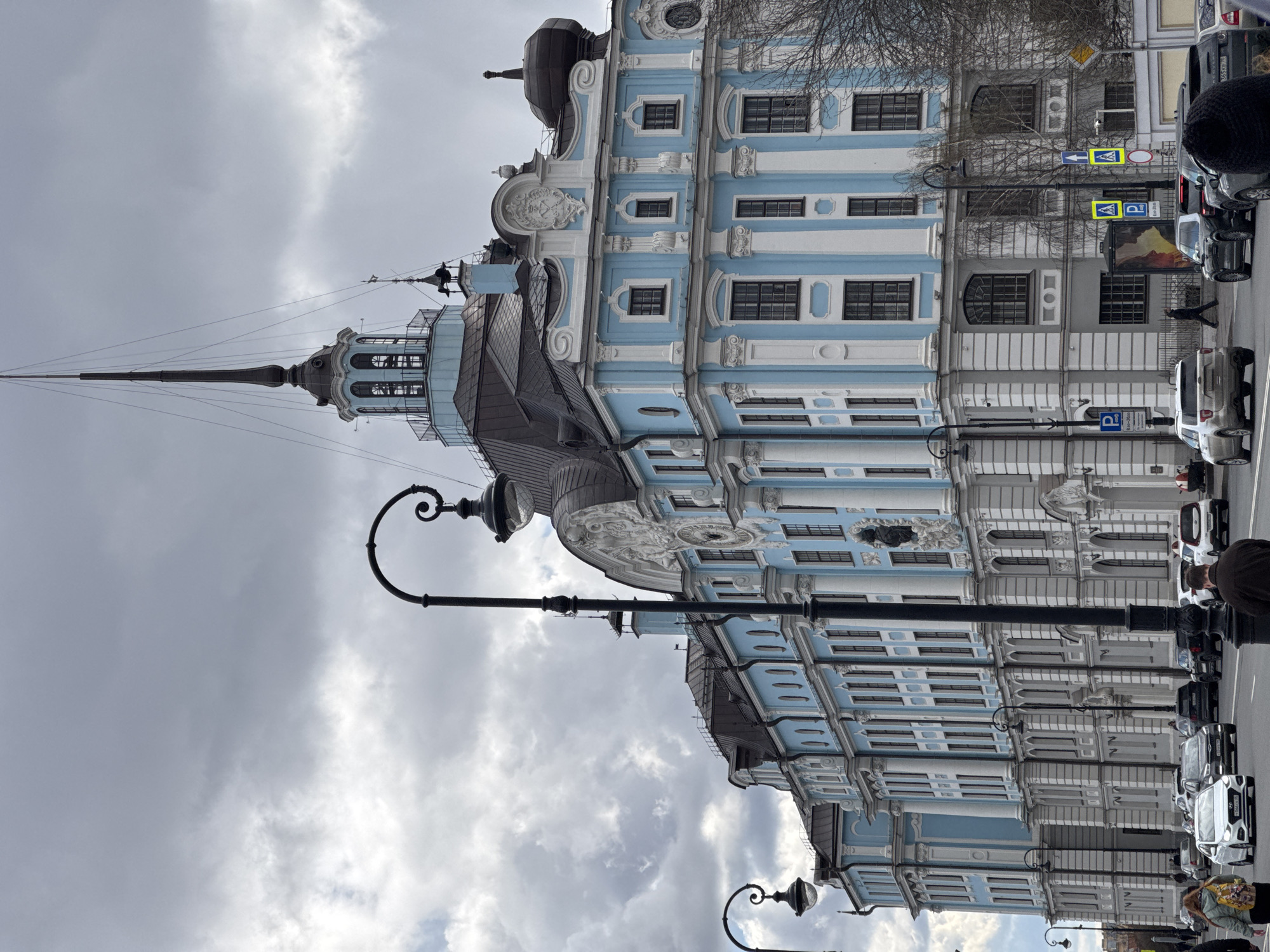 The image shows the Smolny Cathedral in Saint Petersburg, Russia, from a street-level perspective. The cathedral's blue and white facade is prominently displayed, featuring multiple windows, ornate detailing, and a central spire. A portion of the street is visible, with parked cars, street lights, and pedestrian traffic. The sky is overcast with grey clouds.