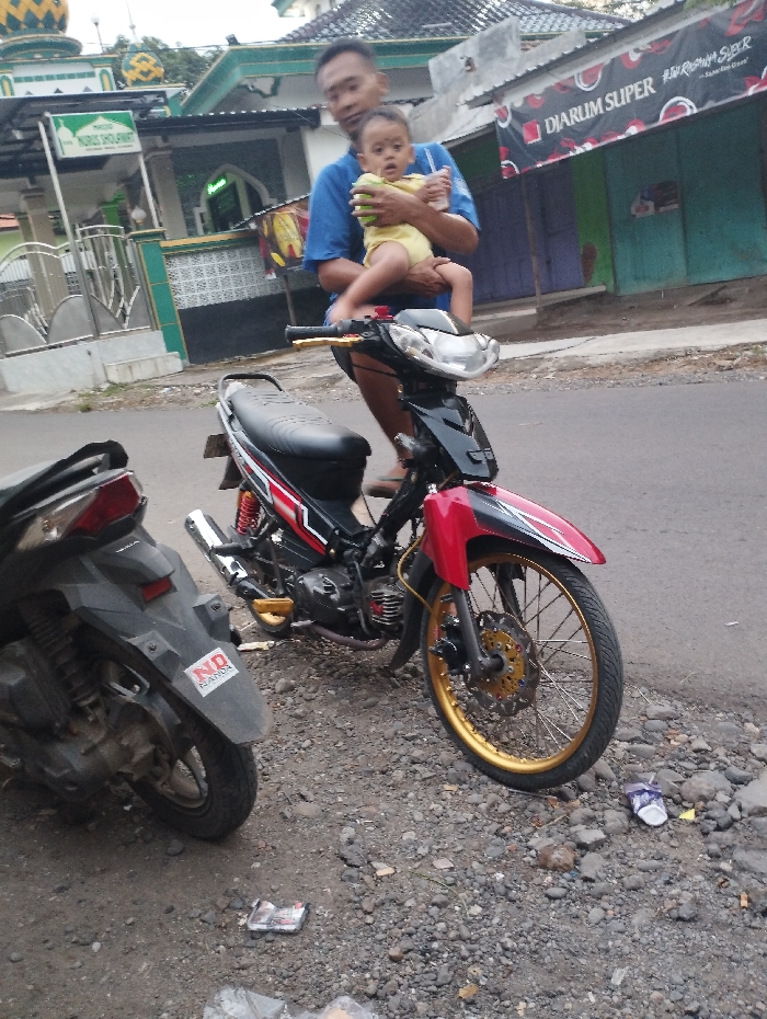 The image captures a man holding a baby, positioned on a red and black motorcycle, parked on a gravel and dirt roadside. The man, wearing a blue t-shirt and shorts, gently holds the baby, who is dressed in a yellow romper and appears to be sitting on the motorcycle's front. Both are looking towards the camera.
The prominent motorcycle is a red and black underbone style, featuring distinctive gold-colored rims, footpegs, and a front brake disc. Partially visible on the left is another dark-colored scooter, whose rear fender has a sticker that reads "ND HONDA". The ground is rugged, consisting of gravel and dirt, with some discarded plastic wrappers and other debris.
In the background, on the left side, there's a building with a green roof and a golden dome, characteristic of a mosque or prayer house. A green sign clearly states "MASJID NURUS SHOUFWANT". To the right, a shop is visible with a red banner across its front, displaying "DIARUM SUPER" and "Djarum Super Mld". The buildings are topped with corrugated metal roofs.
The scene unfolds during the daytime, under soft, diffused lighting, suggesting an overcast sky or a time of day with indirect sunlight. The setting evokes an everyday street or neighborhood scene in a Southeast Asian country, aligning with the location context of Kecamatan Panji, Indonesia.
