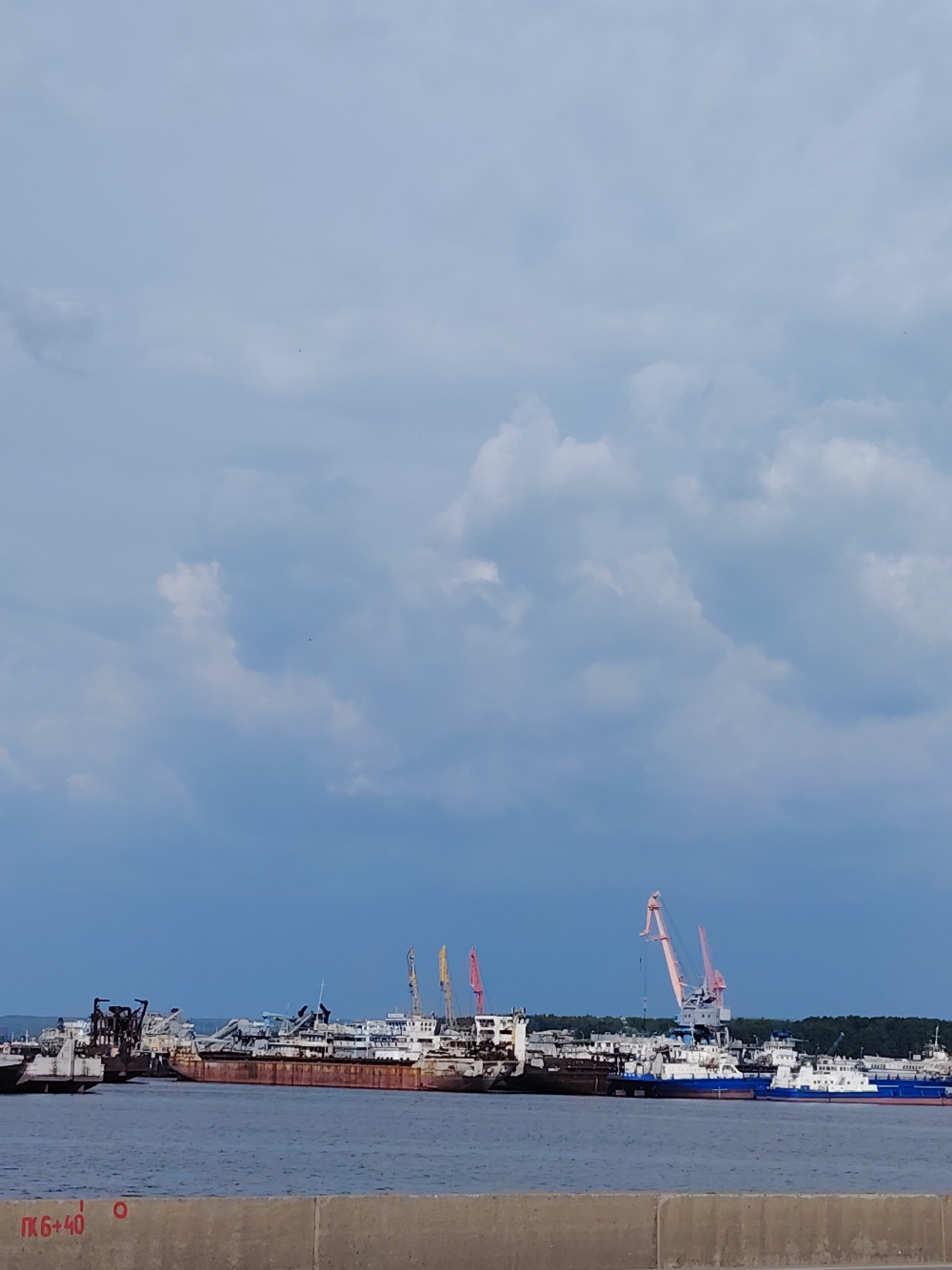 This is a wide shot of a river with several docked ships and cranes. On the left, a large, rusty barge is visible, followed by other ships with dark hulls and white superstructures. Several large construction cranes, some pink and some yellow, stand tall behind the ships. A line of green trees forms a distant backdrop. The sky is a pale blue with scattered white clouds. In the foreground, a concrete barrier with red markings of "ПК 6+40" is present. The image appears to be taken from a waterfront promenade or dock. The location is the Kama River in Chaikovsky, Russia.