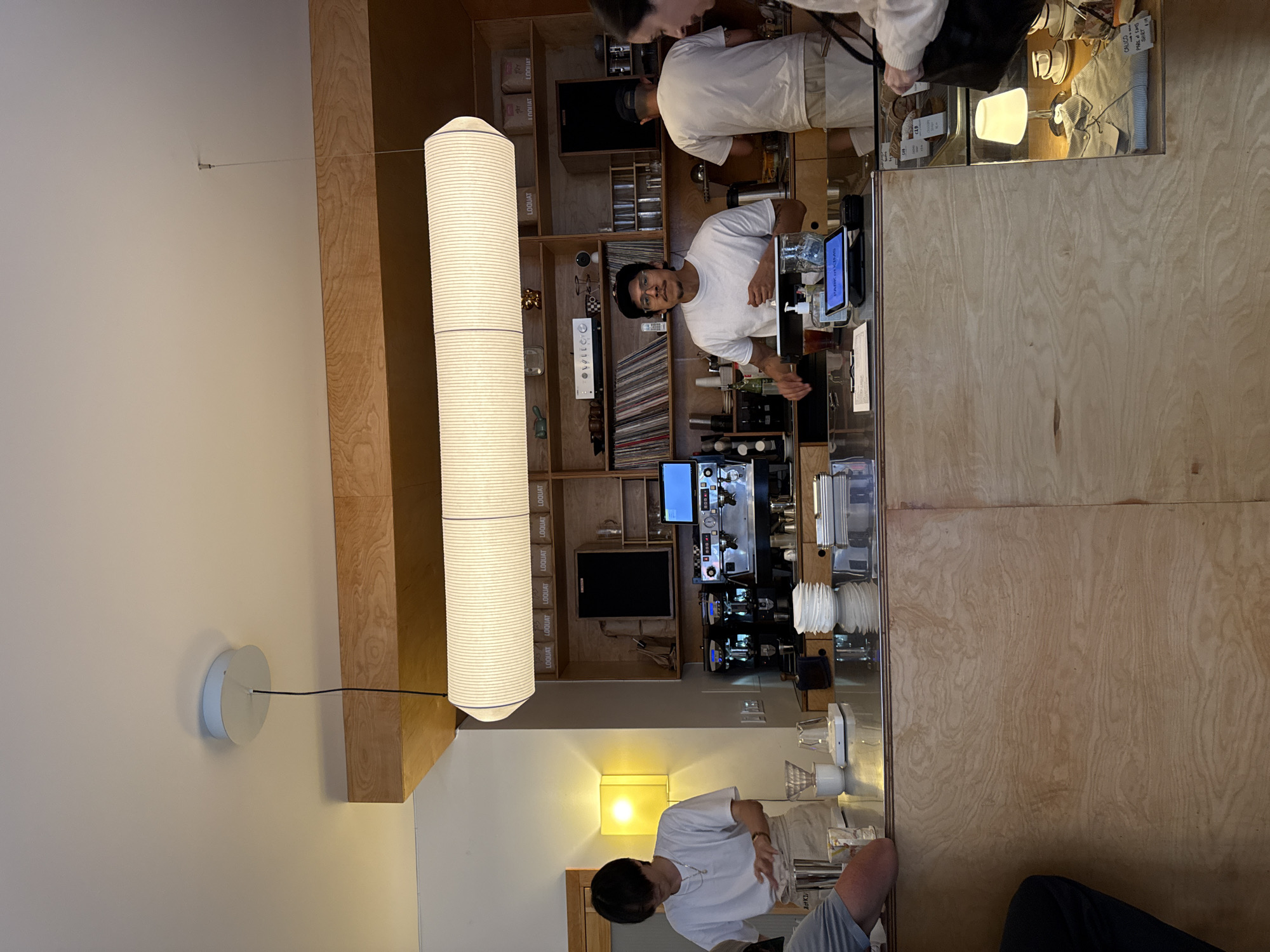 This indoor shot depicts a business environment, potentially a café or coffee shop, located in New York, United States. The setting is a bustling space, evident from the various people present and their interactions. A male, likely a barista, stands behind the counter, his attention seemingly directed towards serving customers. Additional individuals are positioned near the counter, possibly awaiting service. The café's decor includes a bookshelf, adding character to the space. The environment is well-lit. The photograph presents a moment captured inside a local New York business.