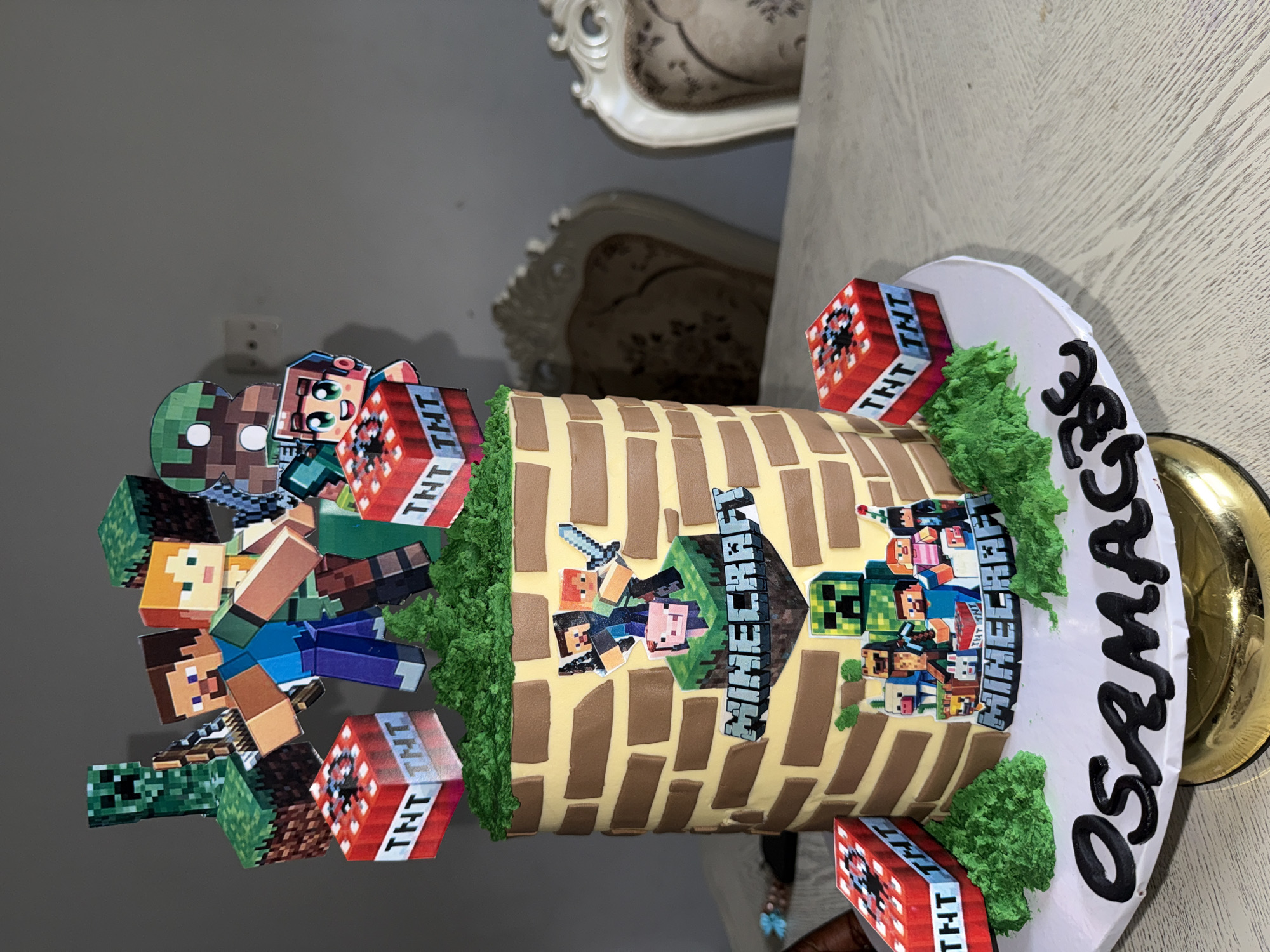 A tilted, high-angle shot features a Minecraft-themed birthday cake on a table, potentially within a residential or event space. The cake has a cylindrical shape and is decorated with brick pattern icing. Minecraft characters, TNT cubes, and green icing resembling grass adorn the top and sides. "OSMACK" is written in black icing on the cake board. The backdrop includes furniture, suggesting an indoor setting. There is no explicit evidence to confirm the image's location within the city of Iddo Tudunwada, Nigeria.