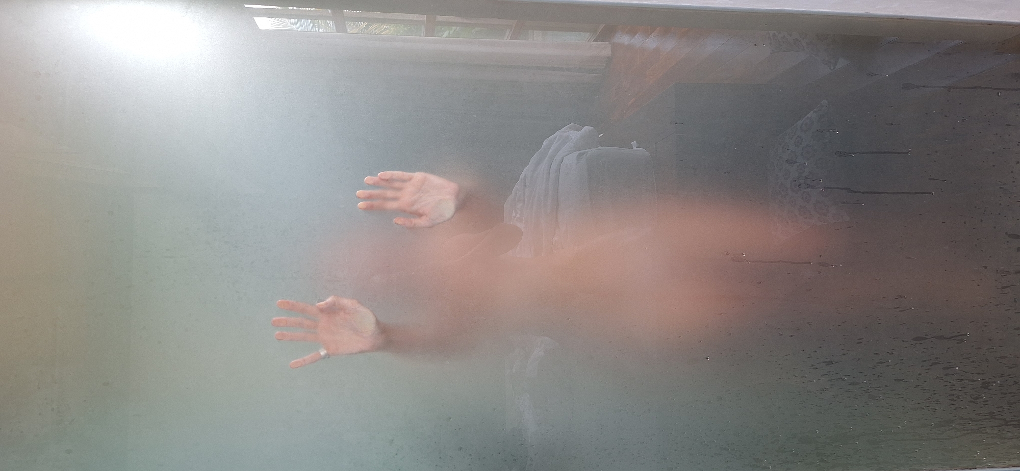 The image shows a person's torso and hands pressed against a steamy glass surface. The person appears to be nude and is facing the viewer. The glass is covered in condensation, obscuring the details of the person's face and body. Two hands are visible on the glass, palms outward, with fingers spread. The left hand has a ring on the index finger. The background is blurred but suggests an indoor setting, possibly a bathroom or shower. A window with curtains is visible on the right side, and a bed with white bedding is seen in the background. The scene is dimly lit, with a bright light source emanating from the right side of the image. There is no information in the image to suggest it is located in Payangan, Indonesia.
