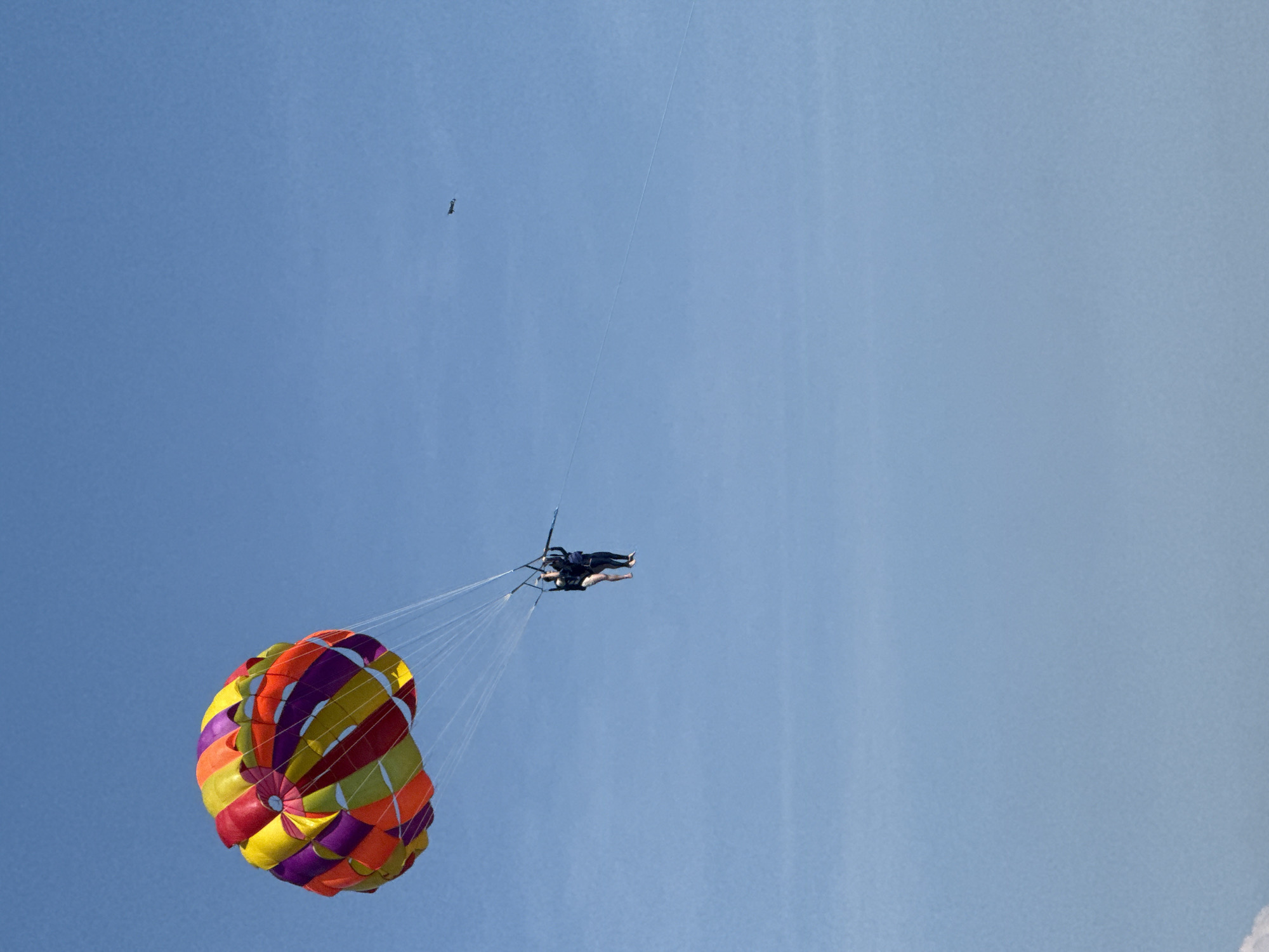Two individuals are parasailing under a vibrant, multi-colored parachute against a clear blue sky. They are suspended from the parachute by ropes, with one person appearing to be behind the other. In the distance, a small aircraft can be seen flying. The scene suggests an outdoor recreational activity taking place on a sunny day. The location is identified as Bac Nha Trang Ward, Vietnam.