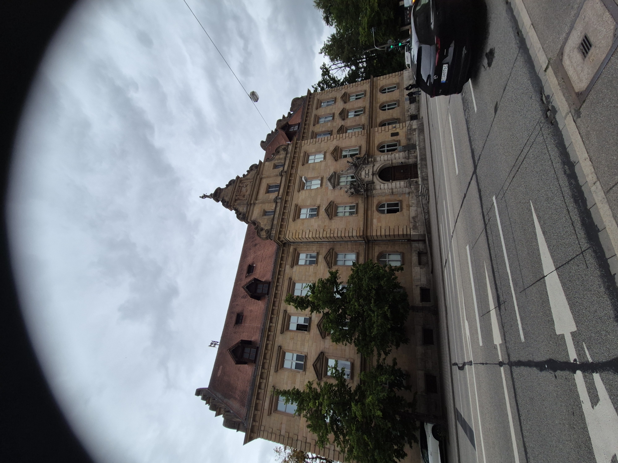 The image depicts a street view of the Old Town Hall (Altes Rathaus) in Regensburg, Germany, under a cloudy sky. The historic building, constructed of light-colored stone, features a prominent red tiled roof with dormer windows and decorative architectural elements, including a statue atop the central gable. The building's facade is punctuated by numerous windows and an ornate main entrance.
In the foreground, a street runs parallel to the building, divided by white lane markings and directional arrows. Two vehicles are visible: a white car parked on the left side of the street near the building and a black SUV parked on the right side of the street, facing away from the camera. The SUV's license plate reads "S-AH976E."
Several mature trees with dense green foliage are positioned between the building and the street. A traffic light is visible on the right side of the image, showing a green signal. The overall environment suggests an urban setting with clear weather, despite the overcast sky. The perspective is slightly distorted, characteristic of a wide-angle lens.