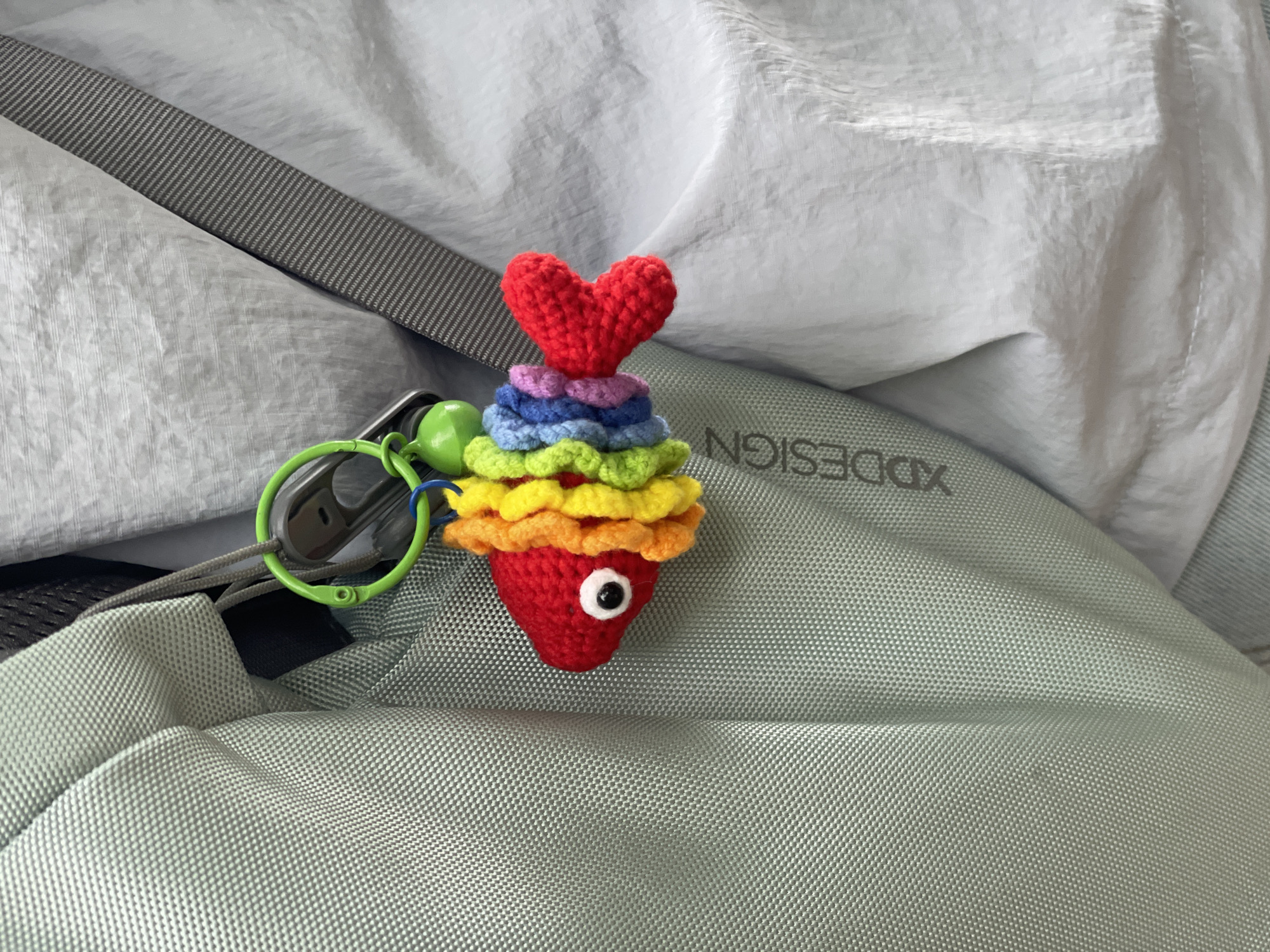 A crocheted keychain resembling a rainbow-layered cupcake with a red heart on top rests on a light green backpack. The keychain has a green ring attached with a green ball and colorful strands. The backpack bears the inscription "XDDESIGN". The backdrop consists of light-colored fabric. The scene appears to be indoors, but the location within Wulingyuan, China cannot be determined from the image.