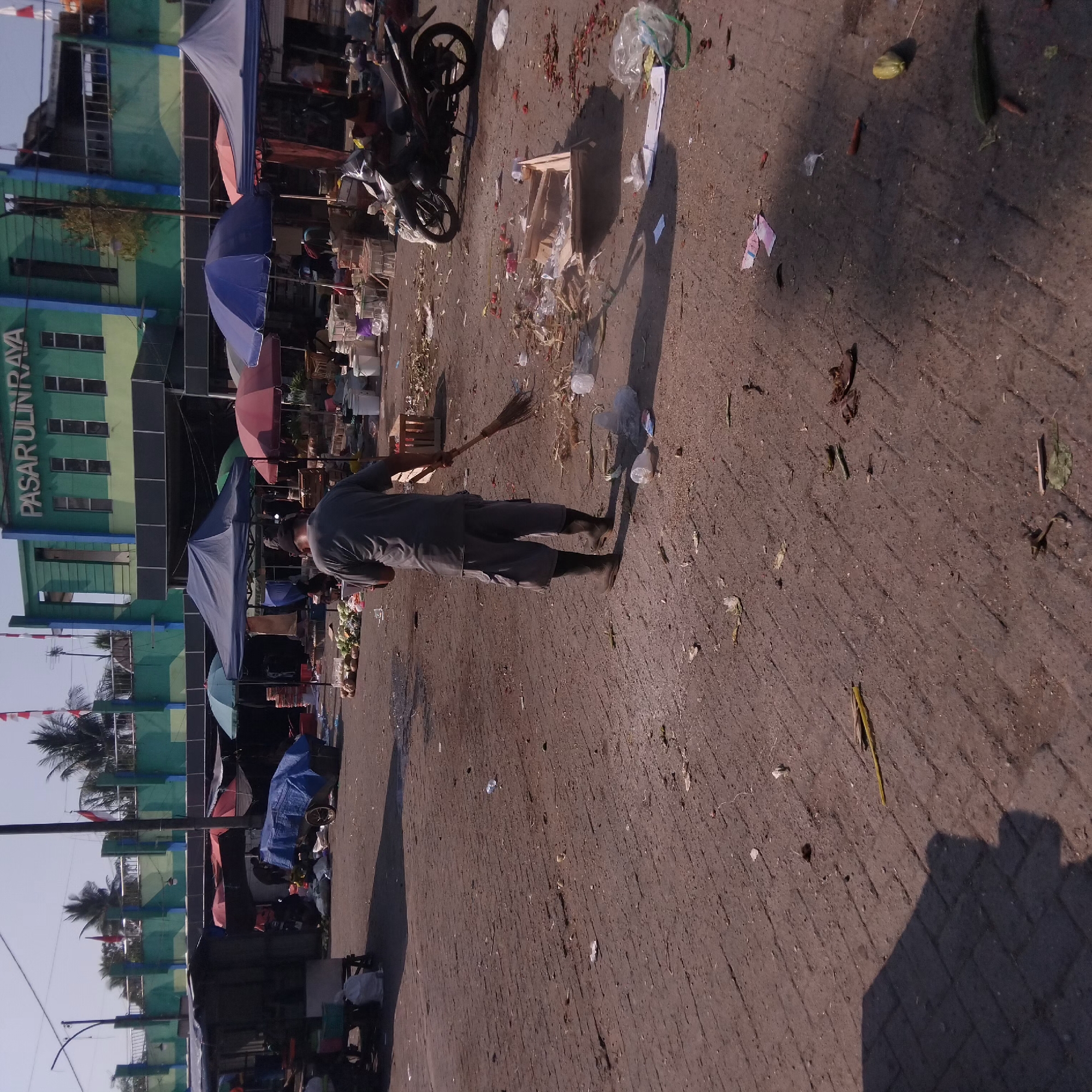 A man is sweeping the ground in front of a market. He is wearing a gray t-shirt, gray shorts, and black boots. He is holding a broom and is actively sweeping. The ground is paved with interlocking bricks and is littered with debris, including leaves, plastic bags, and discarded produce. In the background, there are market stalls with various colorful umbrellas and tarps, and a green building with the words "PASAR ULIN RAYA" written on it, indicating the location is Pasar Ulin Raya in Banjarbaru, Indonesia. A motorcycle is parked on the right side of the image. The sky is clear and blue.