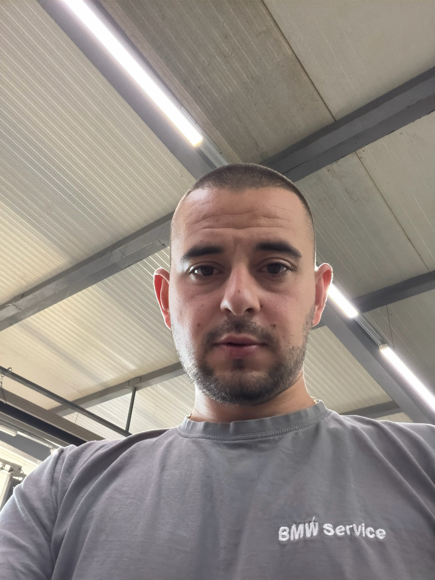 A close-up, low-angle selfie shows a man with a buzz cut and stubble, wearing a grey t-shirt with "BMW Service" printed on it. He is looking directly at the camera. The background consists of a light-colored, ribbed ceiling with fluorescent lights and dark grey support beams, suggesting an indoor industrial or garage setting. The specific location in Burgas, Bulgaria, cannot be determined from the image alone.