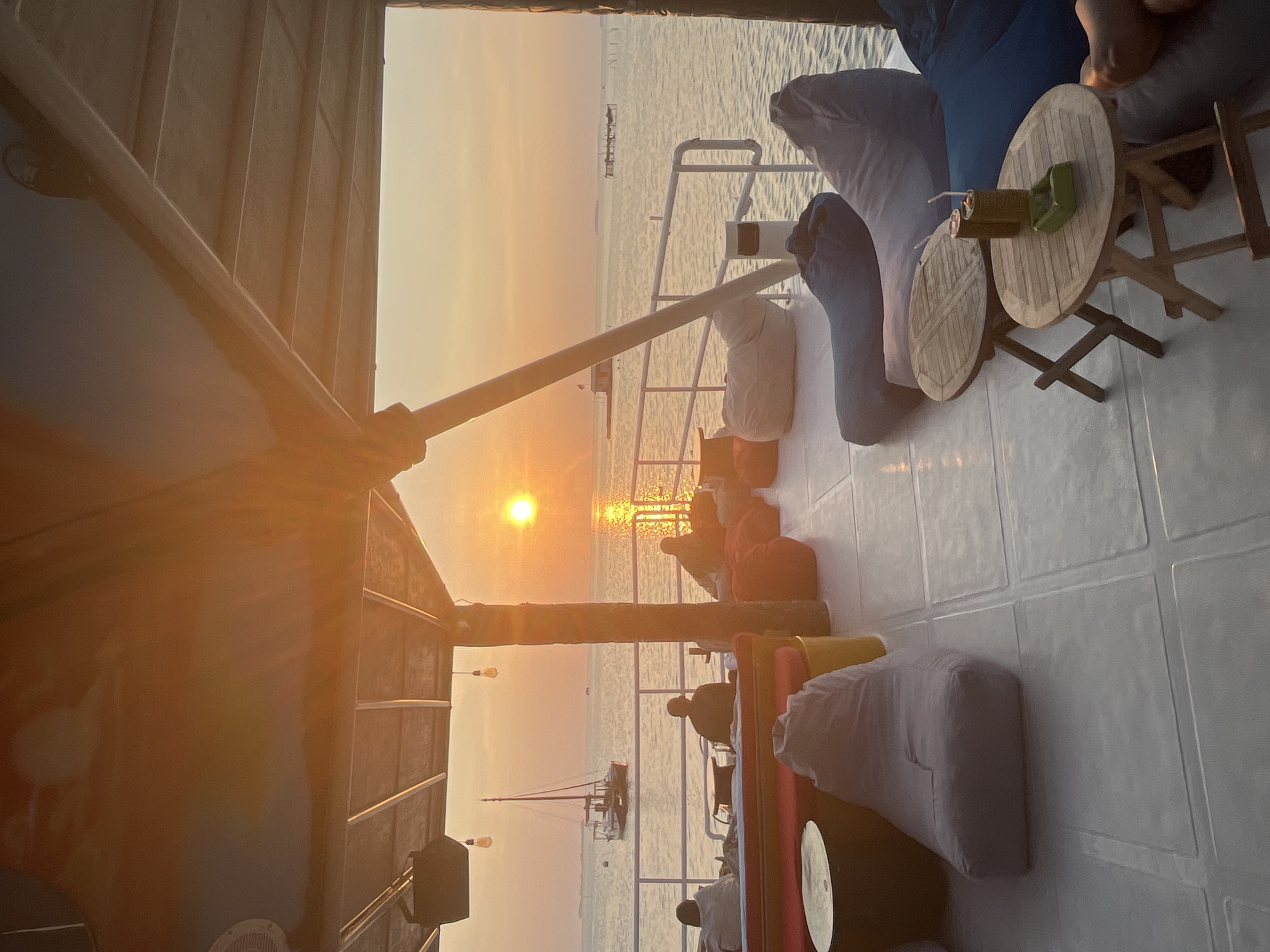 The image captures a serene sunset view from a boat or floating deck in Karimunjawa, Indonesia. Several individuals are seen relaxing on beanbag chairs, facing the tranquil sea. A sailboat is anchored in the distance, adding to the picturesque scenery. The setting sun casts a warm, golden glow across the water and sky, creating a peaceful and inviting atmosphere. The deck is furnished with beanbag chairs, low tables, and some seating with vibrant cushions, suggesting a casual and comfortable outdoor lounge area. The overall scene evokes a sense of relaxation and enjoyment of nature's beauty.