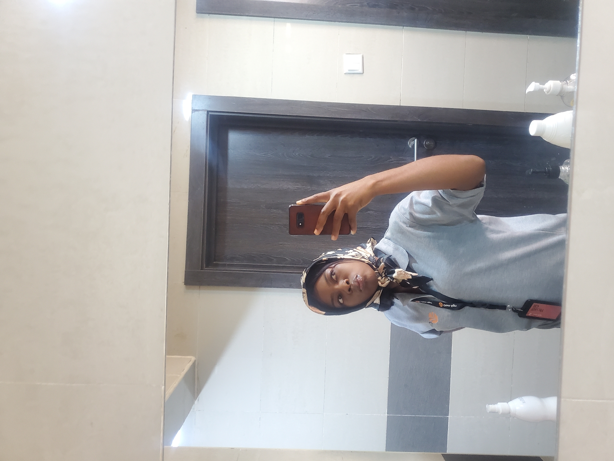 The image captures a rotated mirror selfie of a young woman in an indoor setting. The woman has a medium-dark skin tone and is wearing a light gray long-sleeved shirt and a black and beige patterned headscarf. She holds a dark reddish-brown smartphone in her right hand to take the picture, looking directly towards the camera with glossy lips. A lanyard with an attached ID card is visible around her neck, displaying partially obscured, reversed text that appears to include "THE HOME OF" and "ngà mlû."
The reflected background reveals a clean and modern interior. Behind the woman is a dark brown wooden door with a matching frame. Above the door, the wall is covered in light-colored tiles, featuring a white light switch. To the right of her reflection, two white pump bottles, likely soap or lotion dispensers, are visible on a surface. The lower part of the wall behind her consists of gray tiles with a dark horizontal stripe separating them from the lighter upper section. The setting suggests a public or institutional indoor space, such as an office or facility, located in Gwagwada, Nigeria. No specific time of day or external weather conditions are discernable.