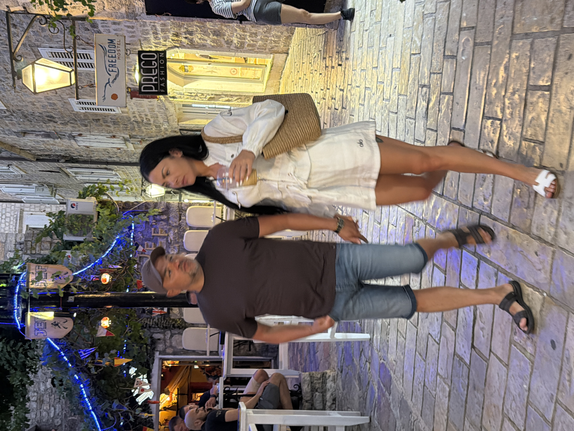 A man and a woman are walking on a cobblestone street in Budva, Montenegro. The man, wearing a brown t-shirt, blue denim shorts, a baseball cap, and dark sandals, walks towards the viewer's left. He has his right hand in his pocket. The woman, wearing a white short-sleeved dress and white sandals, walks slightly ahead of him to his right, holding a drink in her right hand and carrying a straw bag over her left shoulder. She has long dark hair and is looking to her left.
In the background, there is a stone building with shuttered windows and several signs. One sign reads "FREEDOM HOSTEL" with an image of a bird. Another sign says "PREGO FASHION". Blue string lights are draped across a building facade with banners attached. Outdoor seating with white chairs is visible to the left, suggesting a restaurant or cafe. The street is paved with light-colored rectangular stones. The lighting suggests it is evening or dusk.