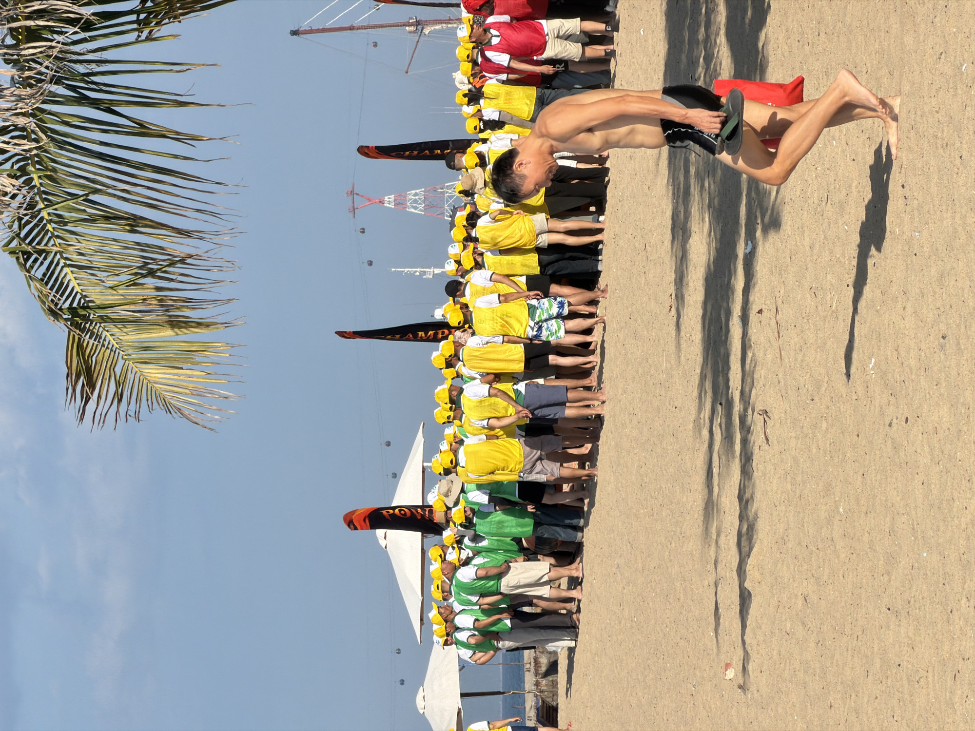 A group of people, predominantly wearing yellow caps and yellow vests over white shirts, are gathered on a sandy beach in Nha Trang Ward, Vietnam. Some are also wearing green vests. They are standing in front of white umbrellas and large orange and black banners. In the foreground, a shirtless man wearing black swim trunks walks away from the camera, carrying a pair of flip-flops in his right hand and a red bag under his arm. The background shows a clear blue sky, a palm frond, a distant red and white tower structure with cables, and what appear to be gondolas or cable cars. The sea is visible behind the umbrellas. The sand is light brown and appears somewhat rippled. Shadows of people and objects are cast on the sand.