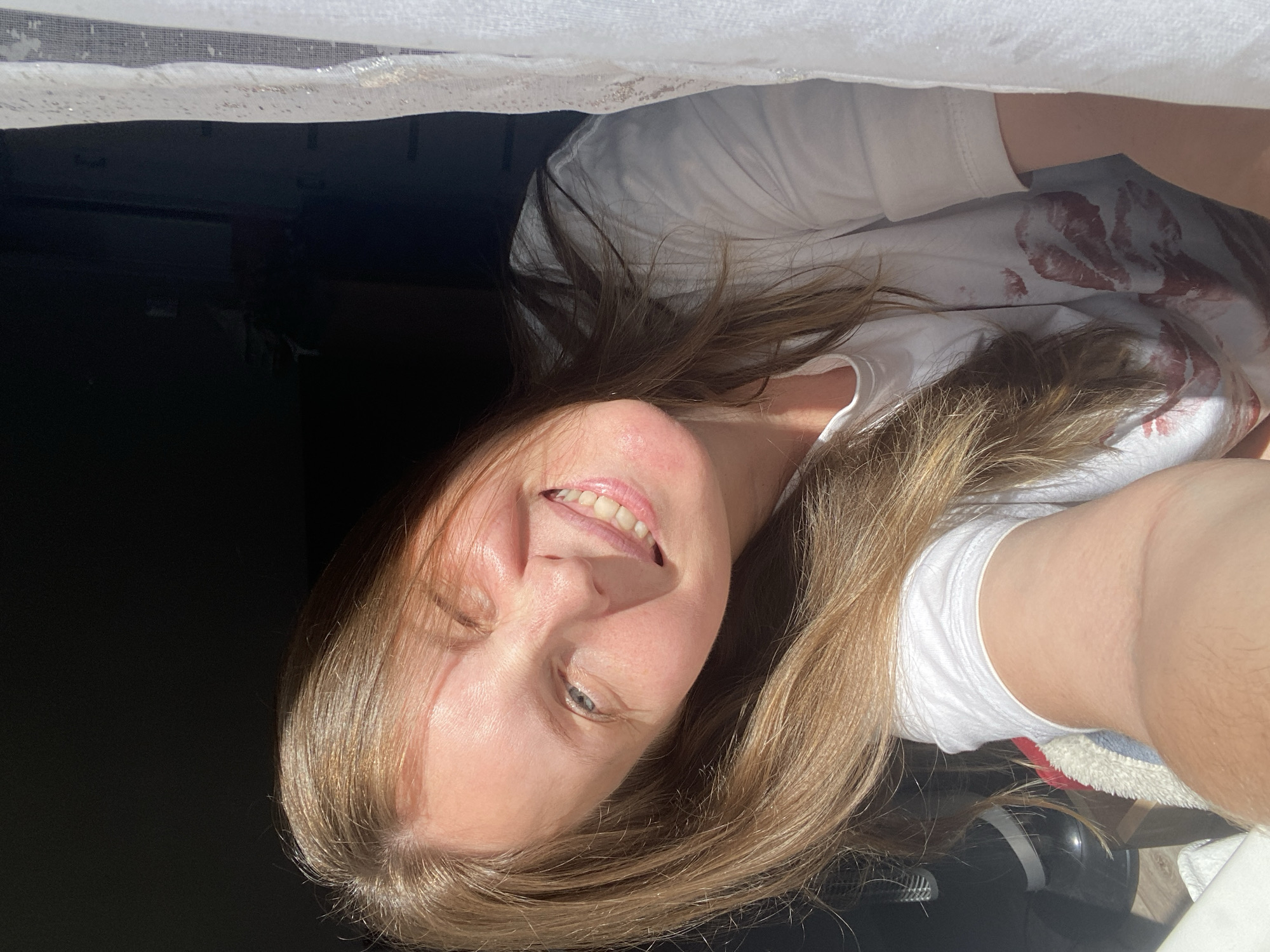 A smiling woman with light brown hair is lying down, looking up at the camera. She is wearing a white t-shirt with multiple red lipstick marks on it. The background is dark and obscured, partially obstructed by what appears to be a white screen. The location is not discernible.