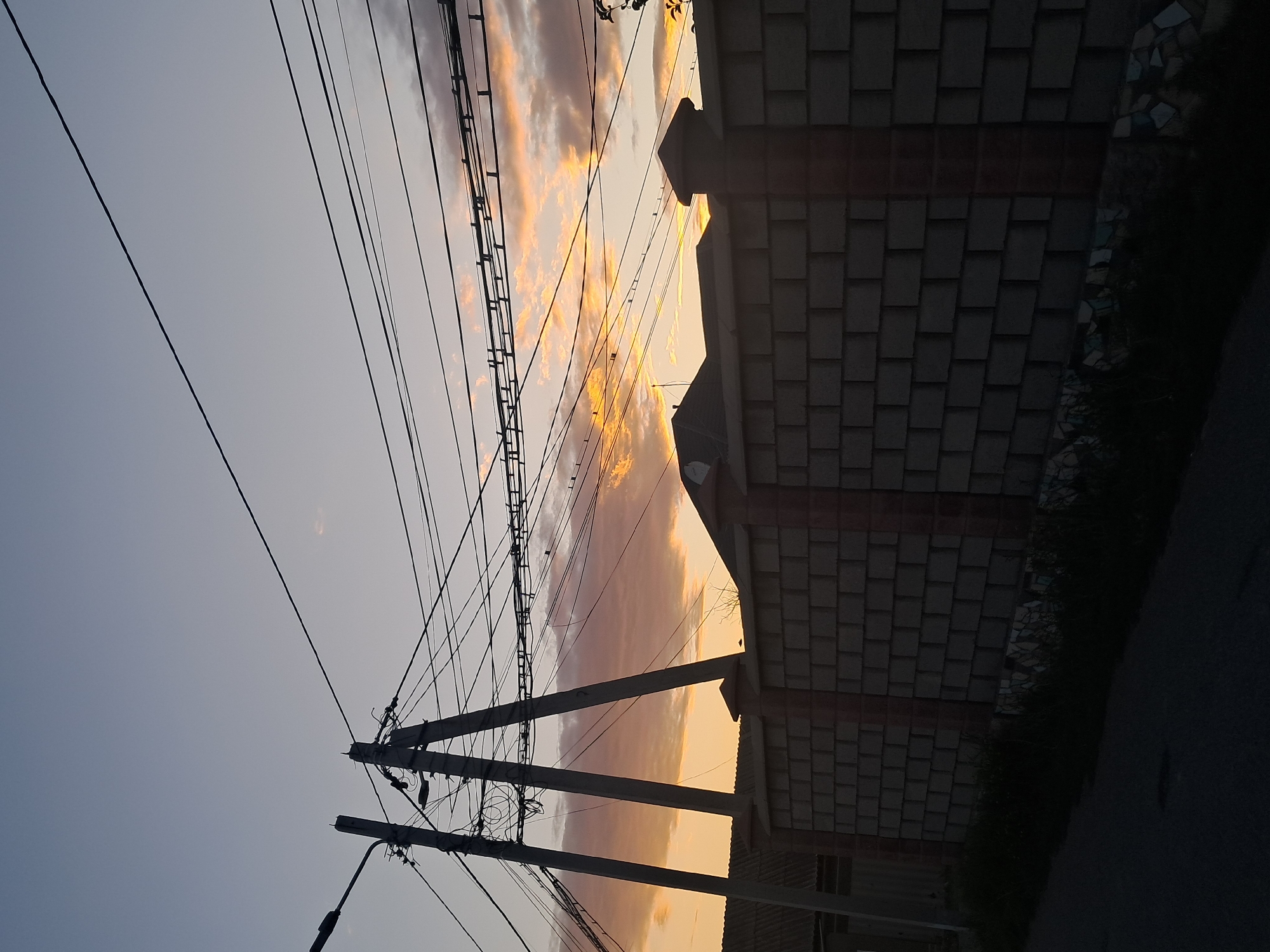 The image depicts an outdoor scene at dusk, characterized by a clear sky with scattered clouds tinged with orange and yellow hues from the setting sun. Overhead, a complex network of power lines crisscrosses the frame, supported by two tall, concrete utility poles. The foreground is dominated by a tall, brick fence, with decorative caps on its pillars. Behind the fence, the silhouettes of rooftops are visible. To the left of the fence, a section of a building with a corrugated roof is partially visible. The ground in front of the fence is a paved surface, possibly a road or sidewalk, with some patches of dark green vegetation growing alongside the fence. The overall impression is of a residential or semi-urban area. The image was taken in Shymkent, Kazakhstan.