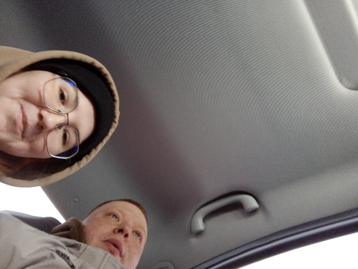 An image taken from a low-angle perspective depicts the interior of a vehicle in Novyi Shagirt, Russia. The vehicle's light grey, ribbed headliner occupies the upper right two-thirds of the frame, featuring a grey overhead grab handle. Two individuals are visible: an adult female in the upper left quadrant, wearing glasses, a dark beanie, and a brown hooded sweatshirt, looking directly towards the camera with a slight smile; and an adult male in the lower left quadrant, with very short hair and fair skin, wearing a light grey jacket, looking upwards and to the right, away from the camera. No direct interaction between the individuals is evident.
