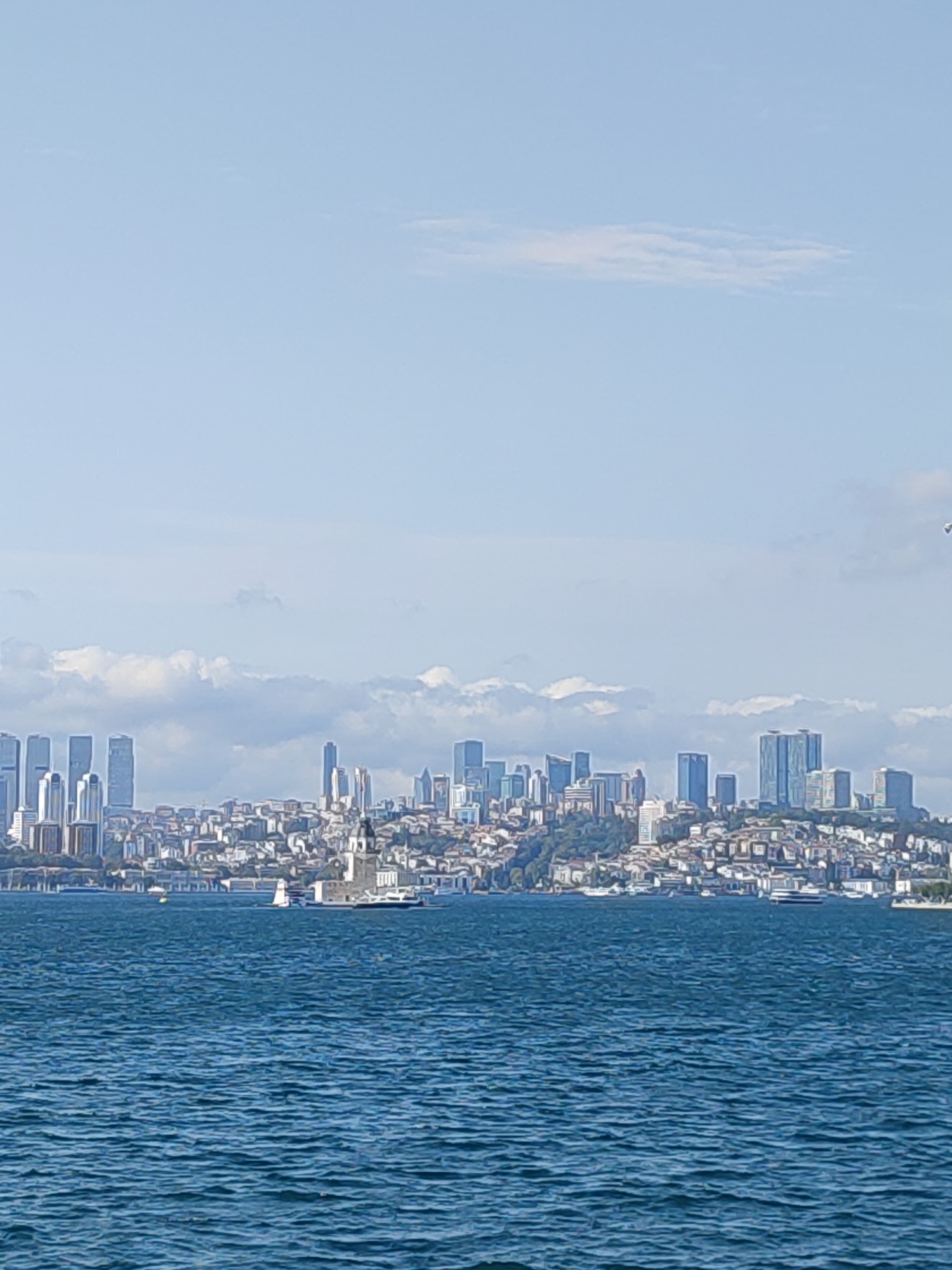 The image shows a wide-angle view of the Bosphorus Strait in Istanbul, Turkey, looking towards the European side. The foreground is dominated by the deep blue, rippling water of the strait, with small waves and reflections. In the midground, the coastline of Istanbul is visible, featuring a dense urban landscape with a mix of old and new buildings. Prominently displayed on a small island in the water is the Maiden's Tower (Kız Kulesi), a historic landmark. Several boats, including a sailboat and what appears to be a ferry, are scattered across the water. The background showcases a modern cityscape with numerous high-rise buildings and skyscrapers, indicative of a bustling metropolis. The sky above is a clear light blue with scattered white clouds, suggesting fair weather.