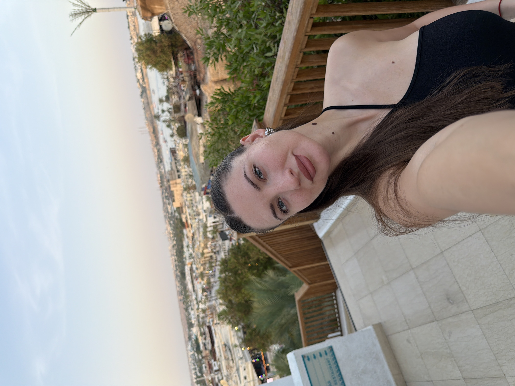 A woman with long, dark hair and light blue eyes takes a selfie outdoors. She is wearing a black halter top and a silver earring on her left ear. A mole is visible on her neck below her ear. Behind her, a railing made of wooden planks separates her from a view of a cityscape and a body of water. The cityscape appears to be in Sharm El Sheikh, Egypt, with buildings and palm trees visible. The sky is clear and pale blue. The ground in the foreground is tiled.