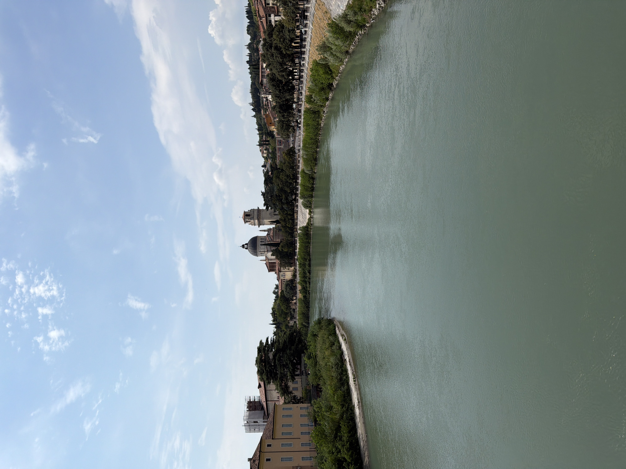 A medium shot captures a wide, greenish river flowing through Verona, Italy, with buildings lining its banks under a bright, partly cloudy sky. The river occupies the majority of the foreground and mid-ground, its surface rippled by a gentle current. On the left bank, a large, pale yellow building with numerous windows is visible, situated amongst lush green vegetation. Further along the left bank, the distinctive domes and spires of a church are prominent, appearing to be the Basilica di San Zeno Maggiore due to its architectural style and location. On the right bank, a tree-lined promenade runs along the river, with a stone embankment supporting it. In the distance, a hillside is covered with buildings and trees, suggesting a city nestled amongst rolling terrain. The overall impression is a scenic urban landscape with a focus on the natural element of the river and the historical architecture of Verona.