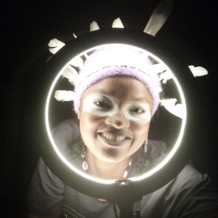 A person with white facial paint and a lavender hat adorned with white feathers is framed by a circular light against a dark background. The individual is smiling, revealing their teeth. They are wearing a necklace and earrings. The lighting accentuates the ring light's glow and creates stark contrasts on the subject's face. The overall effect is theatrical and unconventional.