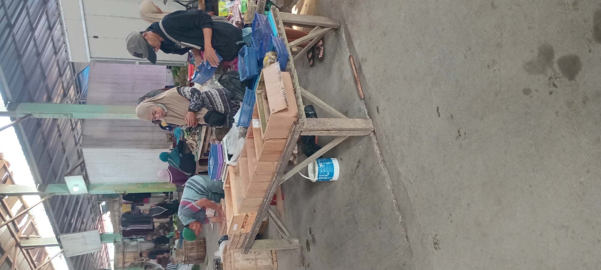 The image depicts an outdoor market scene in Sindangwangi, Indonesia. In the foreground, a wooden table displays several cardboard boxes and blue plastic baskets. A man wearing a green cap and a striped shirt is bent over the table, appearing to arrange items in the boxes. To the right, a woman in a beige hijab and a patterned dress is looking at her phone, standing next to a man in a dark hoodie and a baseball cap who is holding a blue basket. Several other people, some wearing hijabs, are visible in the background, along with stalls and a tiled roof structure. The ground is made of concrete, and there are some discarded items and a bucket on the floor. The overall impression is of a bustling market environment.