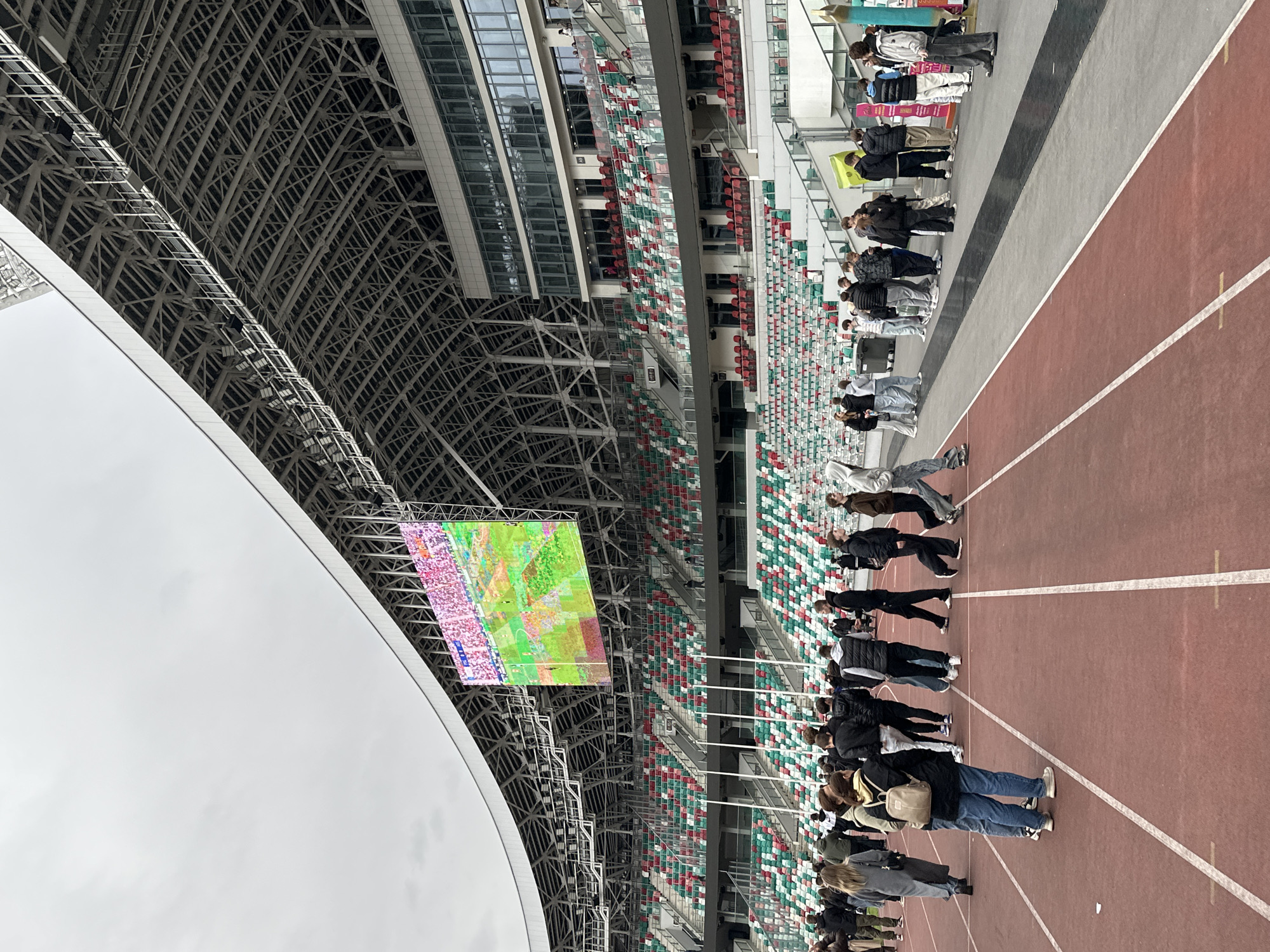 The image depicts a scene inside a stadium in Minsk, Belarus. A group of people are standing near the edge of a running track. The stadium's seating is visible in the background, with a mix of red, white, and green colored seats. Above, a large video screen hangs from the stadium's roof structure. The roof itself consists of a complex network of steel beams. The sky is overcast.