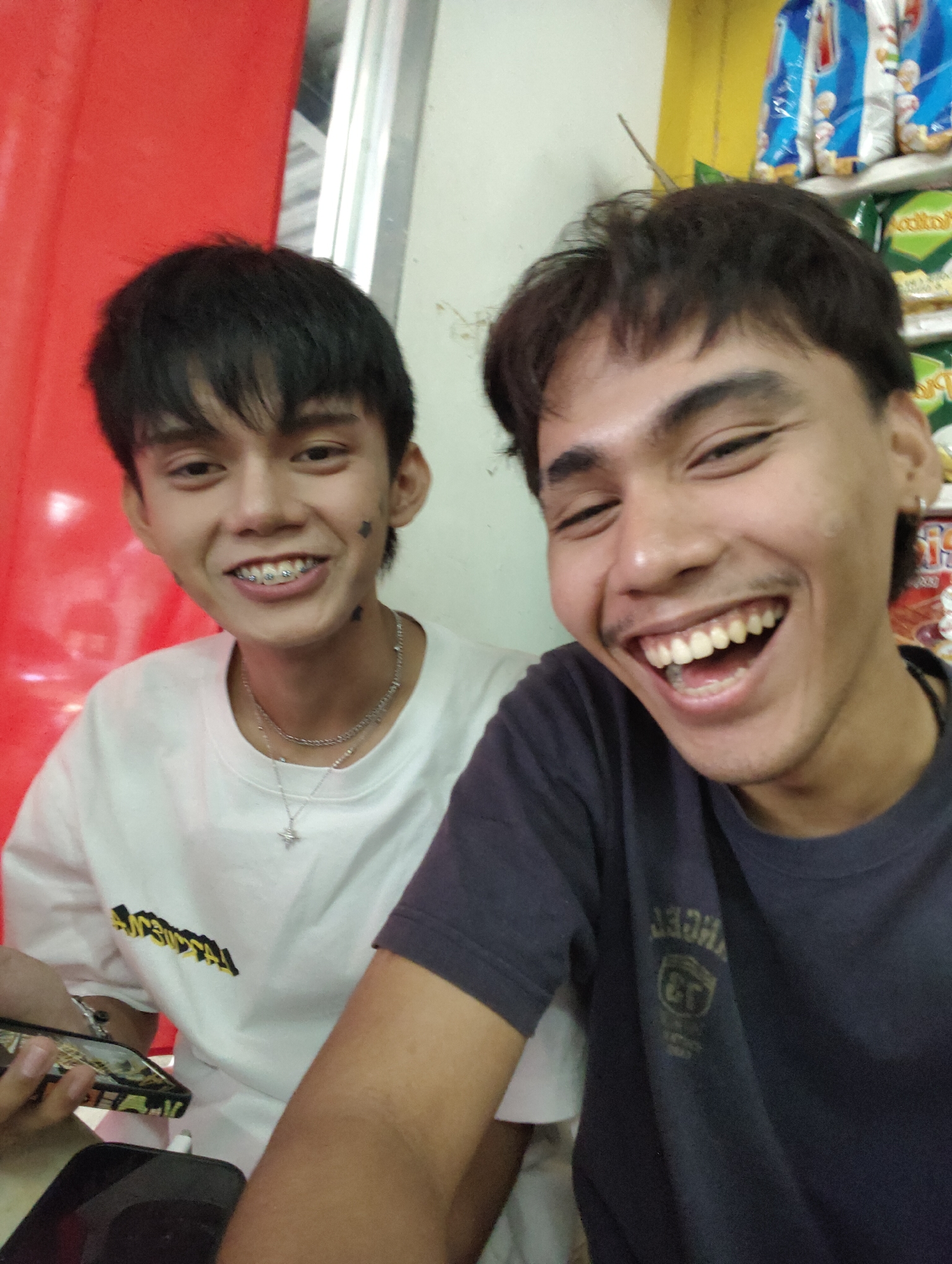 Two young men, appearing to be in their late teens or early twenties, are posing for a selfie. The man on the left has black hair and is wearing a white t-shirt with a yellow graphic and a silver necklace. He has braces and small black markings on his cheeks. The man on the right has black hair and is wearing a dark t-shirt with a faded graphic. Both men are smiling broadly. They are positioned in what appears to be an indoor setting, possibly a convenience store or a small shop, given the shelves of packaged goods visible in the background on the right. The background also shows a red curtain or fabric on the left and a white wall. The lighting suggests an indoor, possibly artificial light source. The image was taken in Davao City, Philippines.
