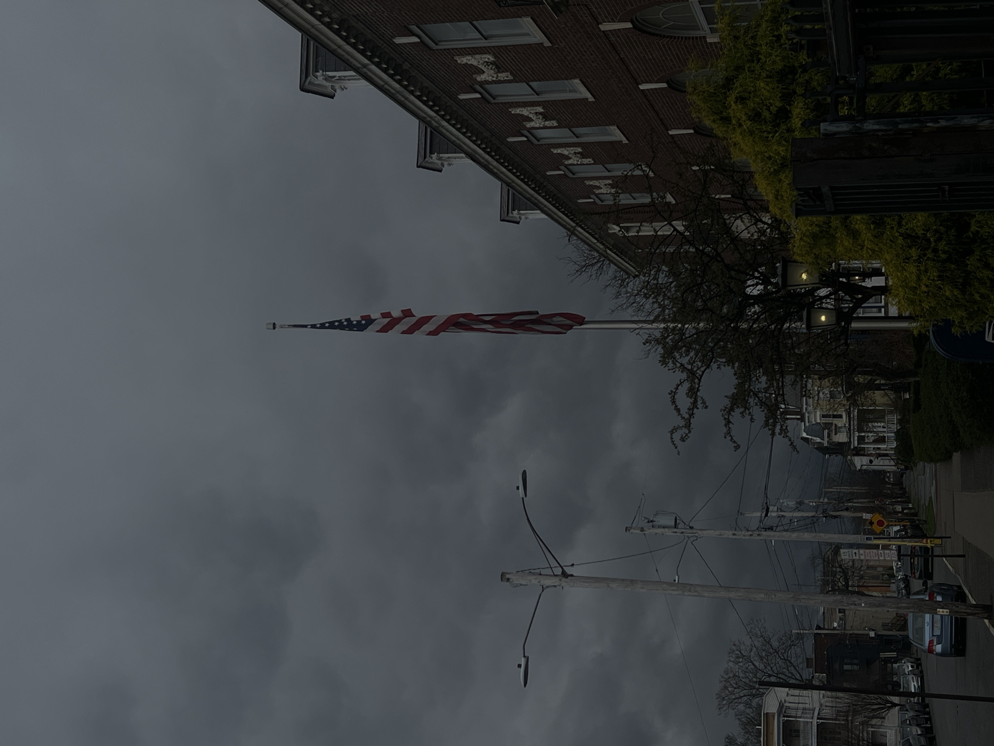 The image depicts a street scene in what appears to be the City of Newburgh, United States of America. The sky is overcast, suggesting a cloudy or stormy weather condition. A large American flag is prominently displayed, extending horizontally from a pole. Adjacent to the flag, a building with a brick facade can be seen. Street lights are present, and along the street, utility poles run parallel, carrying power lines. Cars are parked along the street, and trees line the sidewalks. The image shows a typical urban or suburban streetscape on a gloomy day.