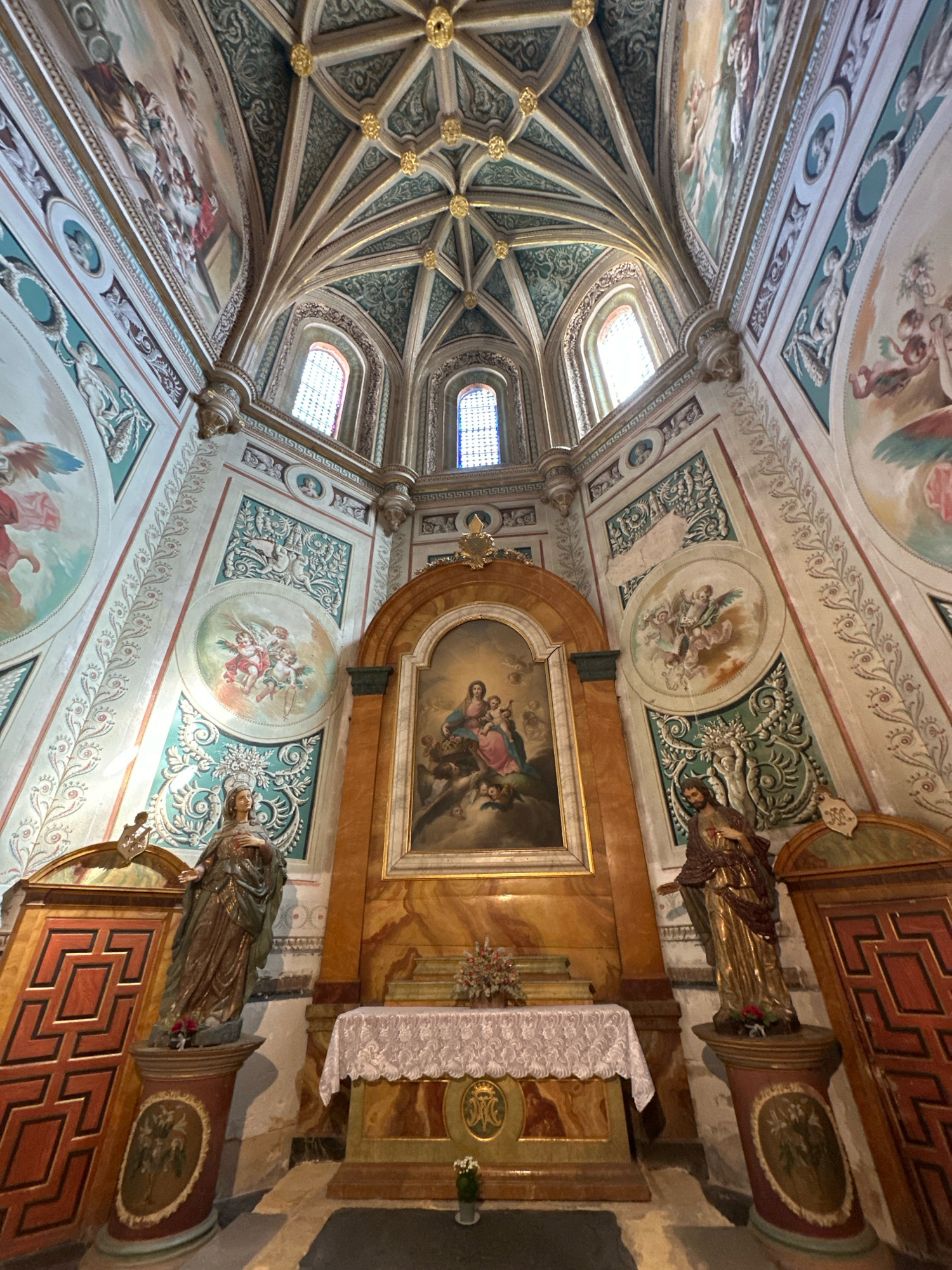 The image shows the interior of a church, likely the Iglesia de San Juan Bautista in La Lastrilla, Spain. The ornate ceiling features Gothic ribbed vaults painted in green and gold. The walls are adorned with frescoes depicting religious scenes, cherubs, and decorative motifs.
In the center of the scene, a large altarpiece features a painting of the Virgin Mary and Child, framed by a golden border. In front of the altarpiece, an altar is covered with a white lace cloth.
To the left of the altar stands a statue of a robed female figure on a pedestal. To the right, a statue of a robed male figure, possibly Christ, also stands on a pedestal. Both pedestals are decorated with circular medallions containing painted scenes.
Flanking the statues are tall, red wooden doors with geometric carvings. The lighting in the church appears to be natural, filtering in through stained-glass windows visible in the upper walls. The overall impression is one of a richly decorated and historically significant religious space.