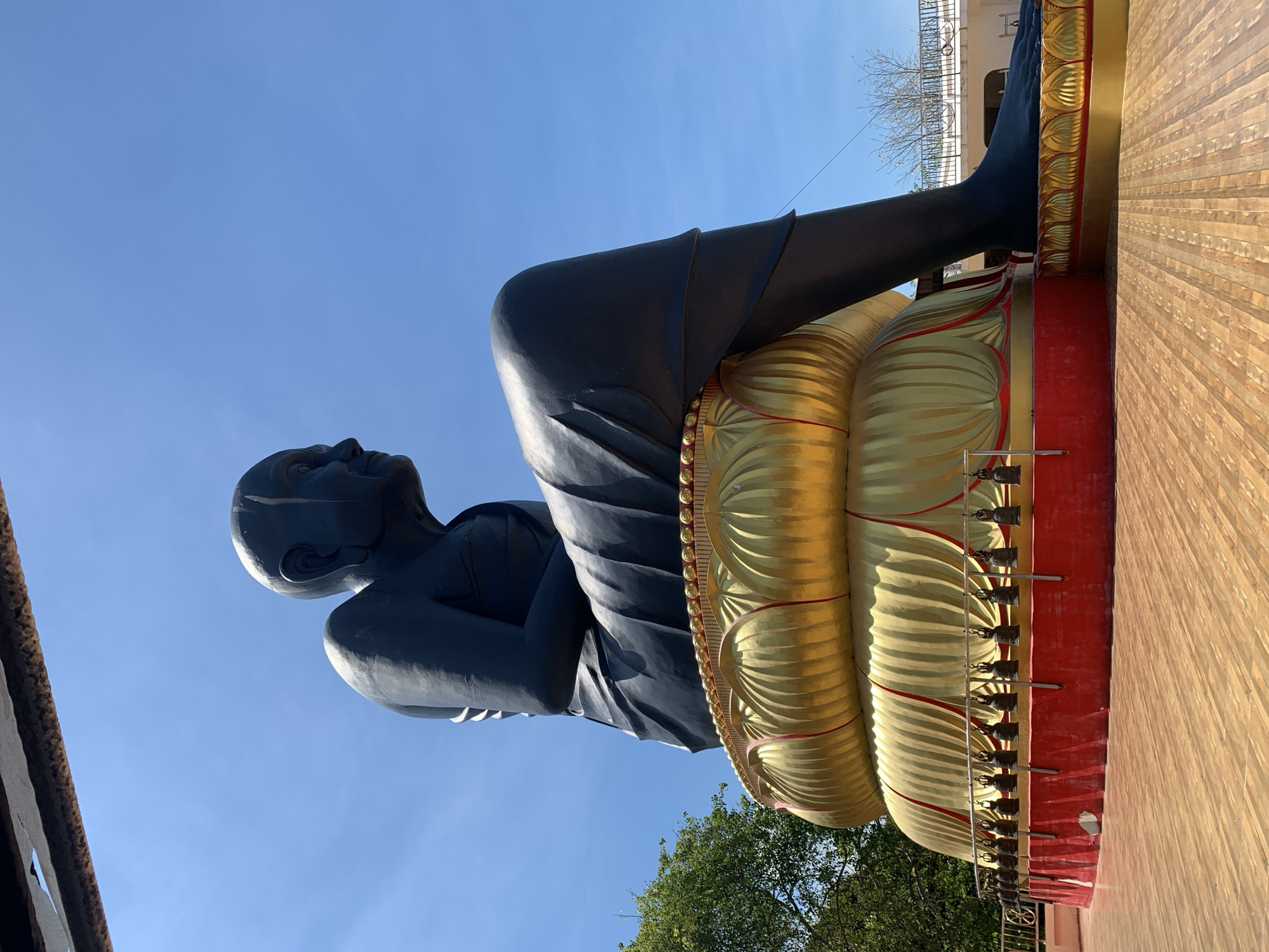 The image shows a large, black statue of a seated figure, likely a Buddhist monk, against a clear blue sky. The statue is depicted in a seated position, with arms crossed in front. It rests upon a multi-layered, golden lotus base bordered with red. Small bells are visible along the lower edge of the statue's base. The statue is located in Baan Bo Phut, Thailand, as indicated by the prompt. The background includes glimpses of surrounding structures and foliage, suggesting an outdoor setting.