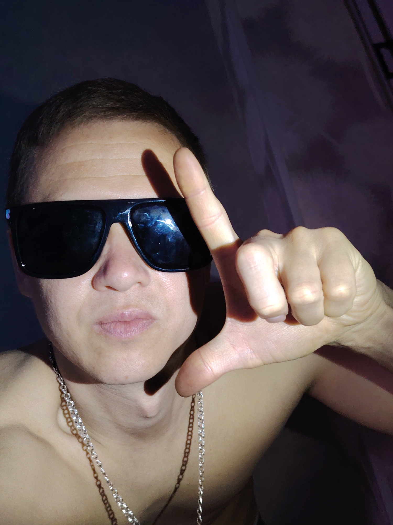 A light-skinned individual with short brown hair is wearing oversized black sunglasses and a silver chain necklace. The person's hand is raised, forming an "L" shape, casting a shadow on their forehead. The background appears to be a dark, blurry, possibly draped fabric. No specific location in Chaikovsky, Russia, is identifiable from the image.