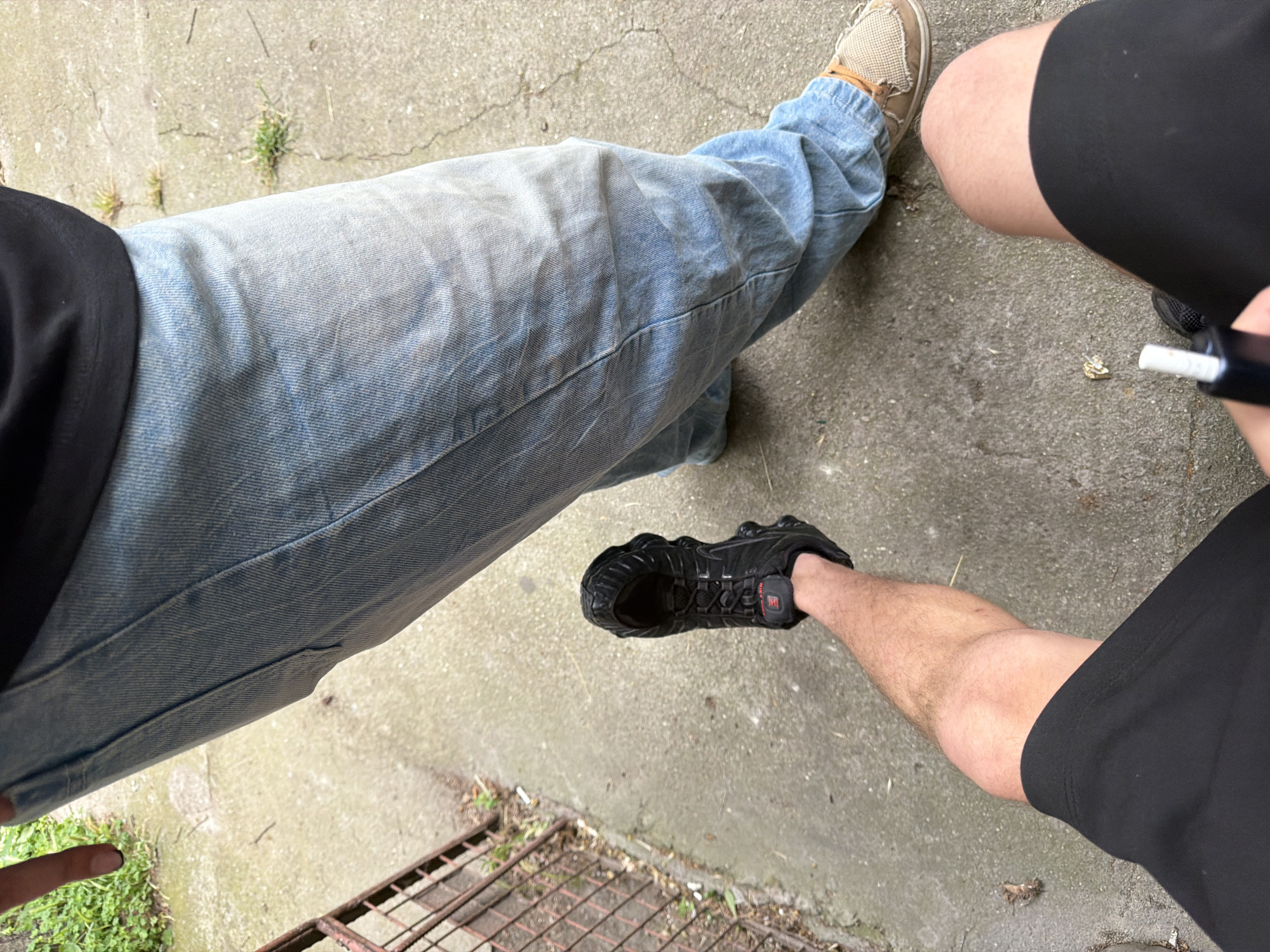 A person, seen from a first-person perspective, is standing on a concrete sidewalk in Plovdiv, Bulgaria. They are wearing black shorts and holding a white cigarette or e-cigarette. In front of them, another person is partially visible, wearing light blue jeans and a black shirt. The second person is wearing black athletic shoes and a light brown shoe with white laces. The sidewalk has patches of green grass growing through cracks. To the left of the frame, a rusty metal fence or gate is visible. The overall environment appears to be outdoors, possibly a residential area or public walkway.