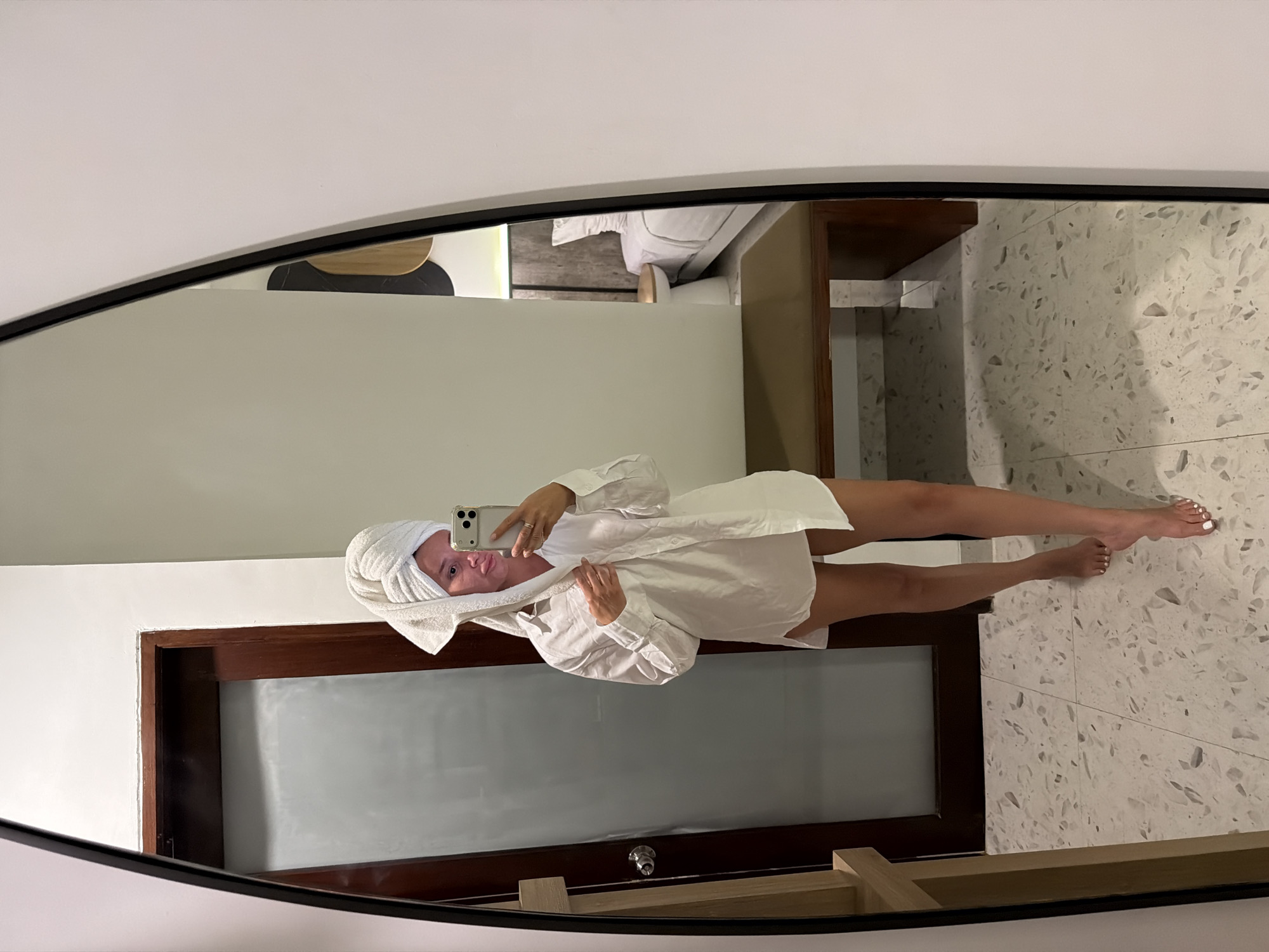A woman with a towel wrapped around her head and wearing an oversized white button-down shirt takes a mirror selfie in what appears to be a hotel room. She is standing on a speckled tiled floor. In the reflection, a door, a wooden bench, and a bed with a headboard are visible. The environment suggests an indoor setting, possibly a bathroom or bedroom.