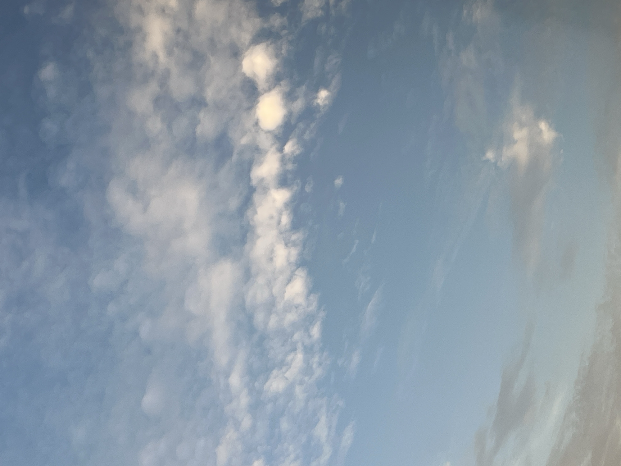 The image depicts a sky filled with scattered white clouds against a pale blue background. The clouds are wispy and appear at various altitudes, with some denser formations and others thinly spread. The lighting suggests it could be daytime, possibly late afternoon or early morning due to the soft quality of light. There are no identifiable landmarks or elements that would place this scene specifically in Warsaw, Poland. The image is solely a depiction of the sky.