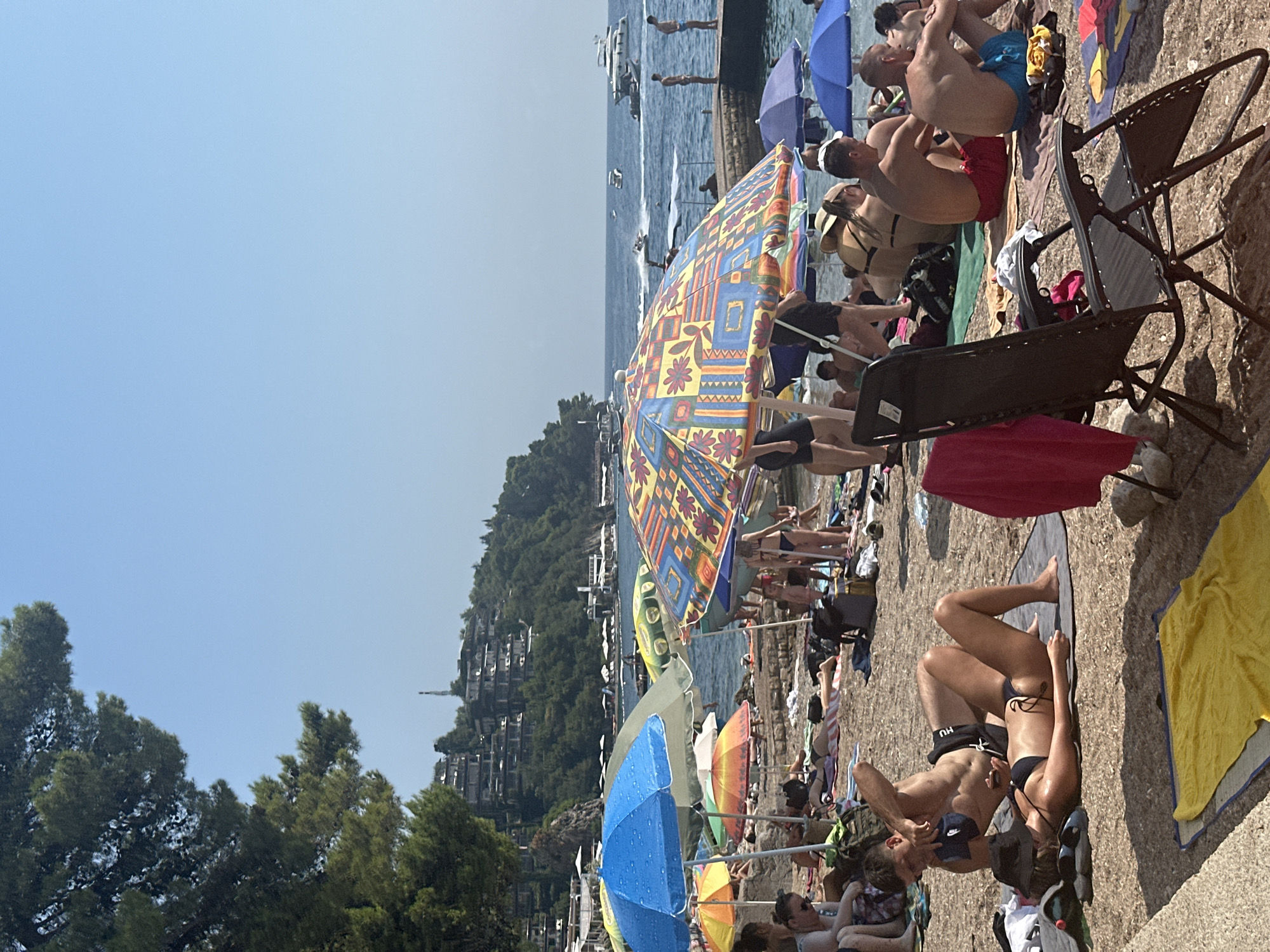 The image captures a sun-drenched beach scene with numerous people relaxing under umbrellas. The beach is composed of small stones, with some areas covered by sand. In the foreground, beach chairs and towels are scattered across the ground, with several individuals sunbathing. A couple is prominently featured in the center, with a man leaning over a woman lying on a towel.
In the middle ground, a variety of colorful umbrellas provide shade to beachgoers. The water is visible behind the beach, with several boats and a jet ski on the surface. A stone pier extends into the water on the right side of the frame, with a few people standing on it.
The background reveals a lush green hillside with buildings constructed on the slope, characteristic of Budva, Montenegro. The architecture of these buildings, with their terraces and stacked design, is typical of the coastal region. The sky is a clear blue, indicating a bright, sunny day. The overall scene depicts a busy and lively beach resort atmosphere.