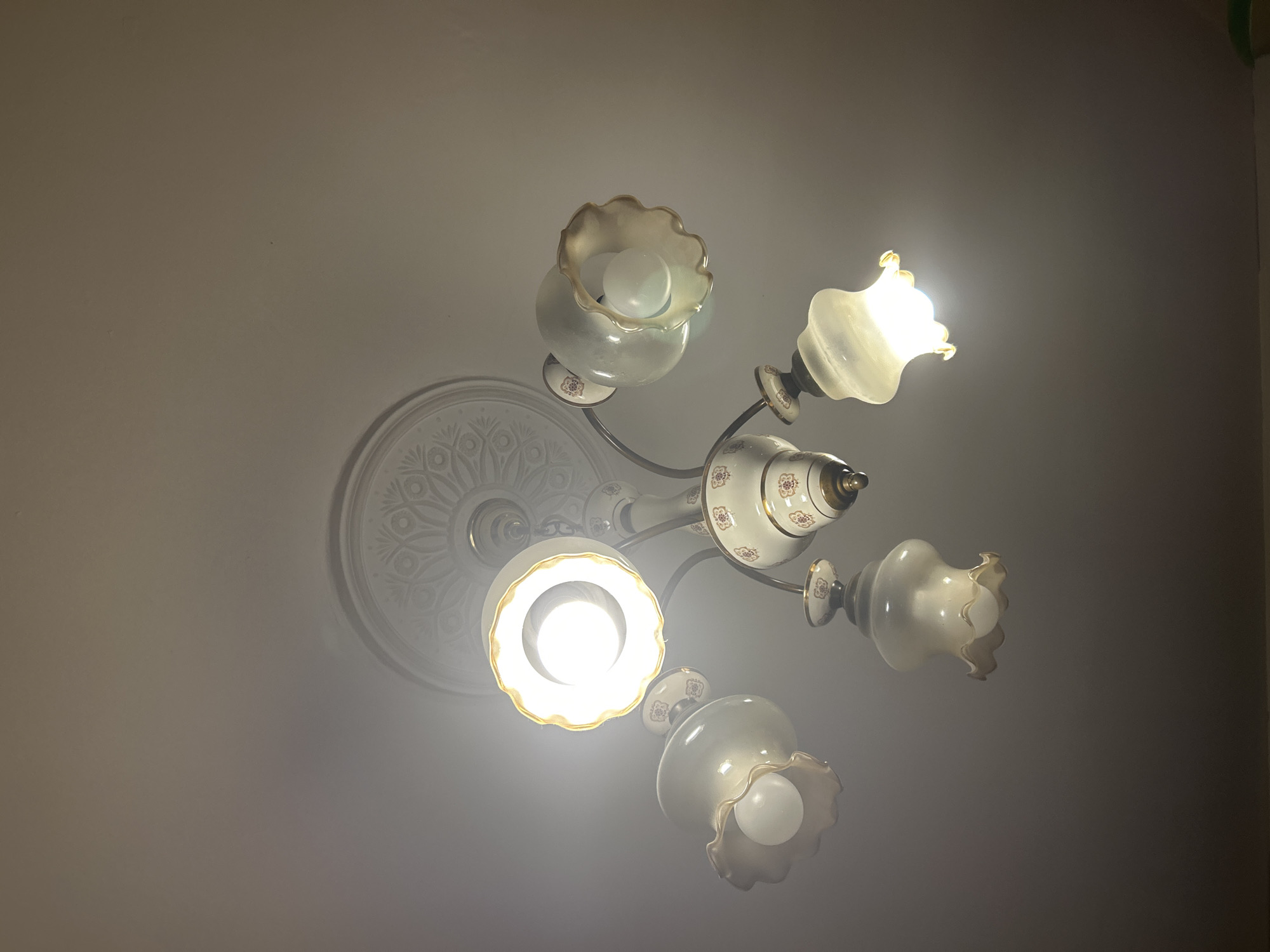 The image shows a chandelier with five lights attached to a decorative ceiling medallion. The chandelier has a white base with gold accents and floral detailing in red and gold. The light fixtures are also white with gold rims and a scalloped edge. Four of the lights are illuminated, with one being significantly brighter than the others. The ceiling appears to be textured. The location is not specified.