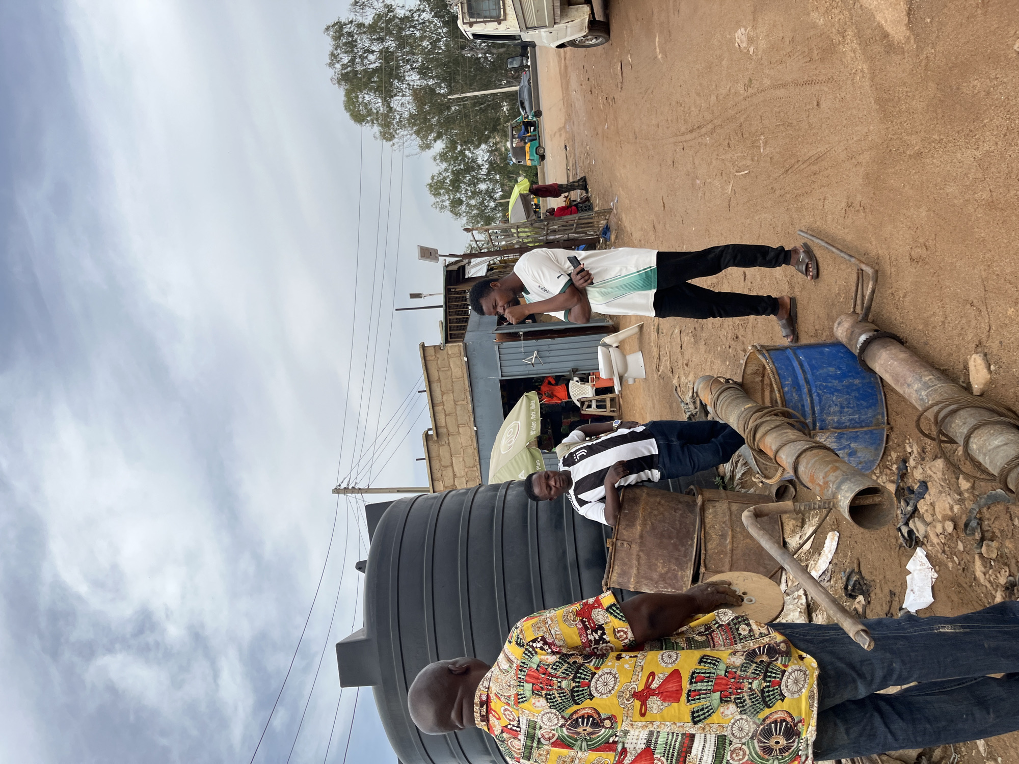 Three men are present in the scene, two of whom are standing and one seated. The man in the foreground is wearing a colorful patterned shirt with yellow as the dominant color, and blue jeans. He is facing away from the camera. Another man is standing in the mid-ground, wearing a white t-shirt with green stripes and black pants. He is holding a phone in his hand and looking down at it. The third man is also standing in the mid-ground, wearing a black and white striped t-shirt and dark pants. He is leaning on a rusty metal object and looking towards the camera.
The scene appears to be outdoors in a dusty, possibly rural or semi-urban area. There are several objects scattered around, including a large black water tank, a blue barrel, and various metal pipes and scrap materials. In the background, there is a building with a corrugated iron roof and a small truck. The ground is dirt and uneven. The sky is overcast, suggesting cloudy weather.
The activity taking place is not entirely clear, but the individuals appear to be engaged in casual conversation or individual activities. The man with the phone might be making a call or browsing. The presence of the water tank and pipes suggests that this might be a place related to water storage, distribution, or repair.
Notable details include the diverse clothing of the men and the mix of new and discarded items in the environment. The lighting is diffused due to the cloud cover. The location cues suggest Nigeria. There is no visible text in the image that can be clearly deciphered. The overall mood appears relaxed and ordinary.