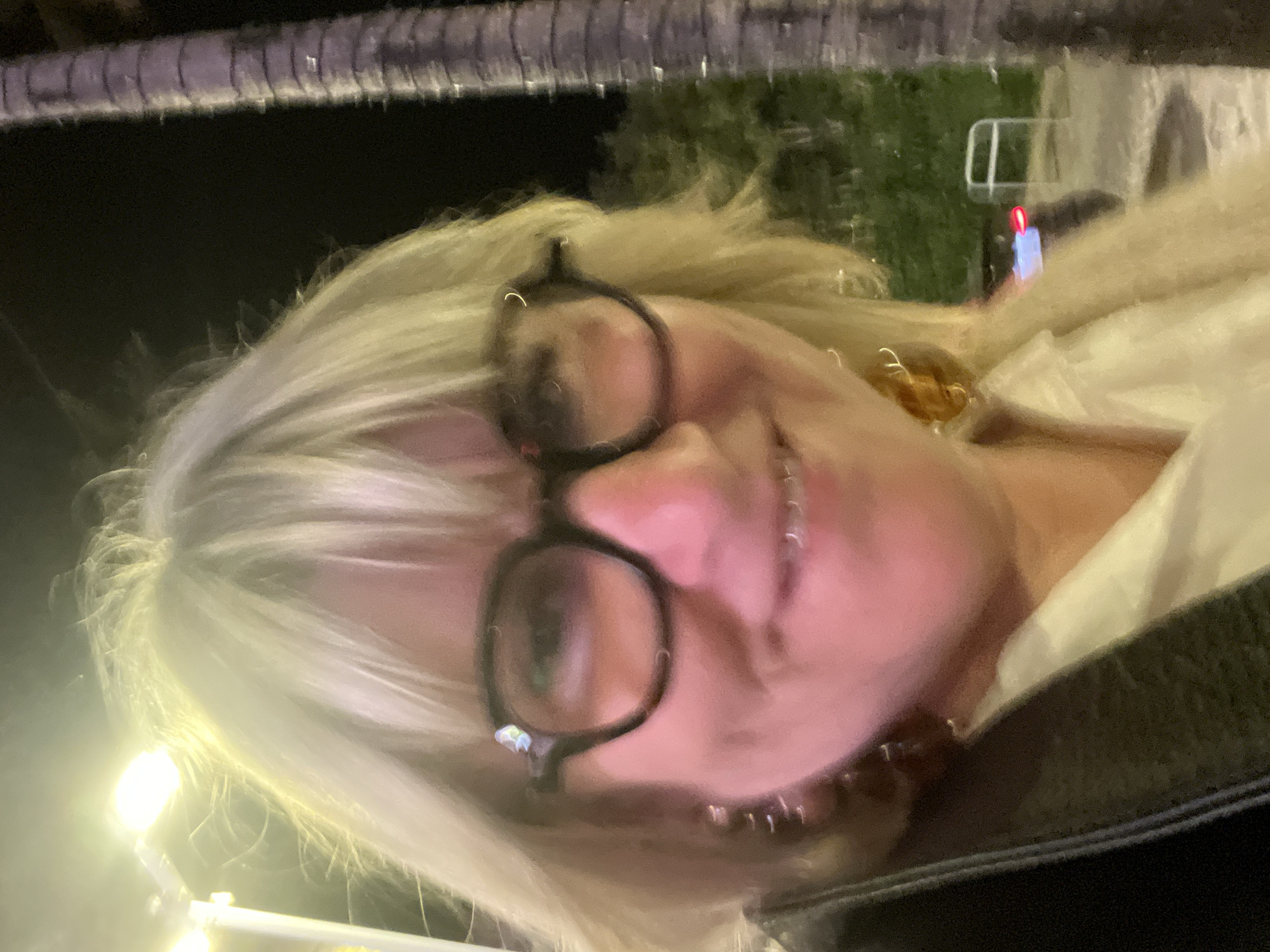The image shows a woman with blonde hair and glasses smiling at the camera. She is wearing a white shirt and a black jacket. The background is blurry, but it appears to be a street scene at night. A light post is visible in the lower left corner. In the background, there is a structure with a red light, and some trees. Given the context, this is likely in the city of Cavalaire-sur-Mer, France.