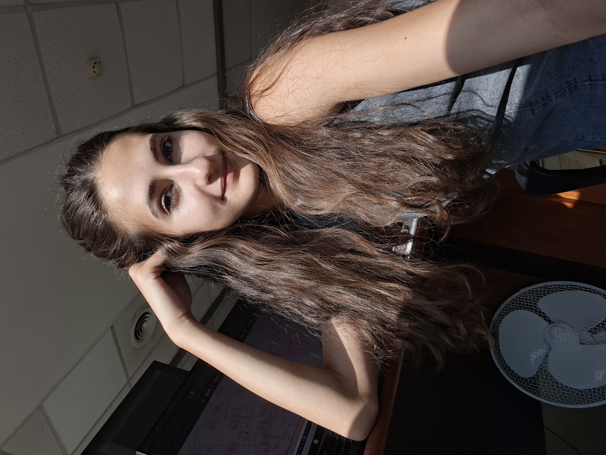 In a dimly lit room, a young woman with long, wavy brown hair leans her right arm on a dark surface, her hand resting on her head. She is wearing a gray tank top. Sunlight streams in from the right, casting shadows across her arm and hair, and illuminating her face. A white fan with a metal grate is visible in the lower left corner of the image, next to a dark desk or counter. In the background, a computer monitor displays what appears to be a diagram or chart, and a drop ceiling with light fixtures and a circular vent is visible overhead. The overall scene suggests an indoor workspace or office environment.