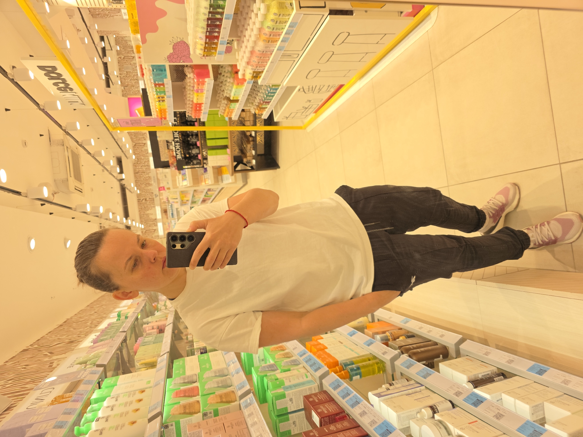 A person with dark hair pulled back into a bun, wearing a white t-shirt and dark pants, stands in a brightly lit cosmetics store, taking a mirror selfie with a black smartphone. The person's left hand is in their pocket. Red string bracelets are visible on their left wrist. The store shelves are stocked with various beauty products, displayed in colorful packaging. A prominent yellow shelf divider with text "DOROGOMIR" is visible in the background. The floor is tiled in a light beige color. The lighting is warm, creating a golden hue over the scene. The store appears to be in Rostov-on-Don, Russia, based on the signage.