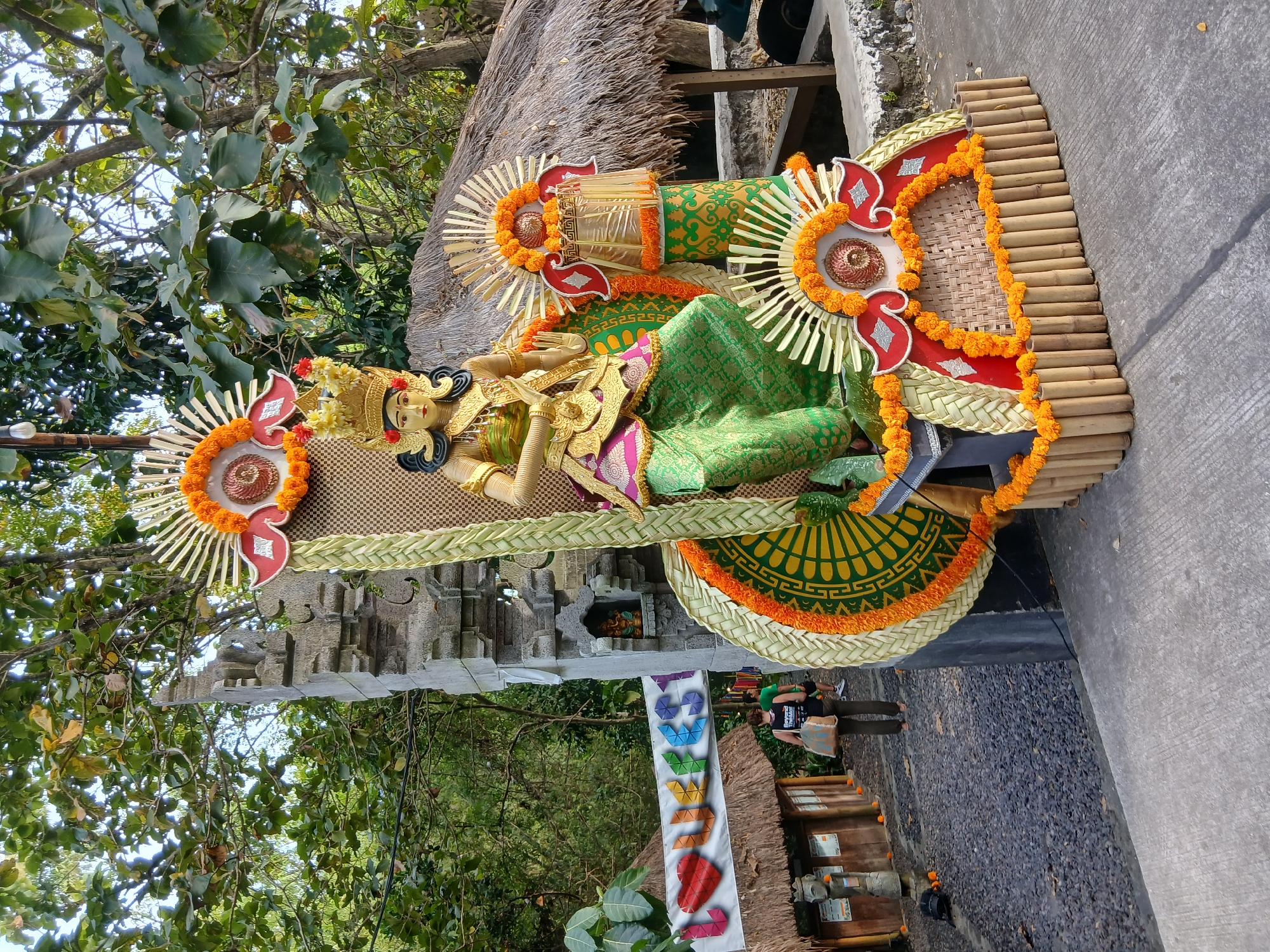 A statue of a Balinese dancer is on display outdoors, adorned with vibrant decorations of marigolds, woven palm leaves, and bamboo. The statue is gilded, wearing a golden crown, a gold and pink top, and a green patterned skirt. It sits on a platform decorated with bamboo and more marigold garlands. Behind the statue is a stone temple gate with intricate carvings, and further back, a thatched-roof hut. A banner with colorful letters spelling "LOVEST" is visible to the left. The ground is covered with dark gravel and concrete. Two individuals are walking in the background, one carrying a bag. The scene is set amidst lush green trees, suggesting a natural or cultural setting.
