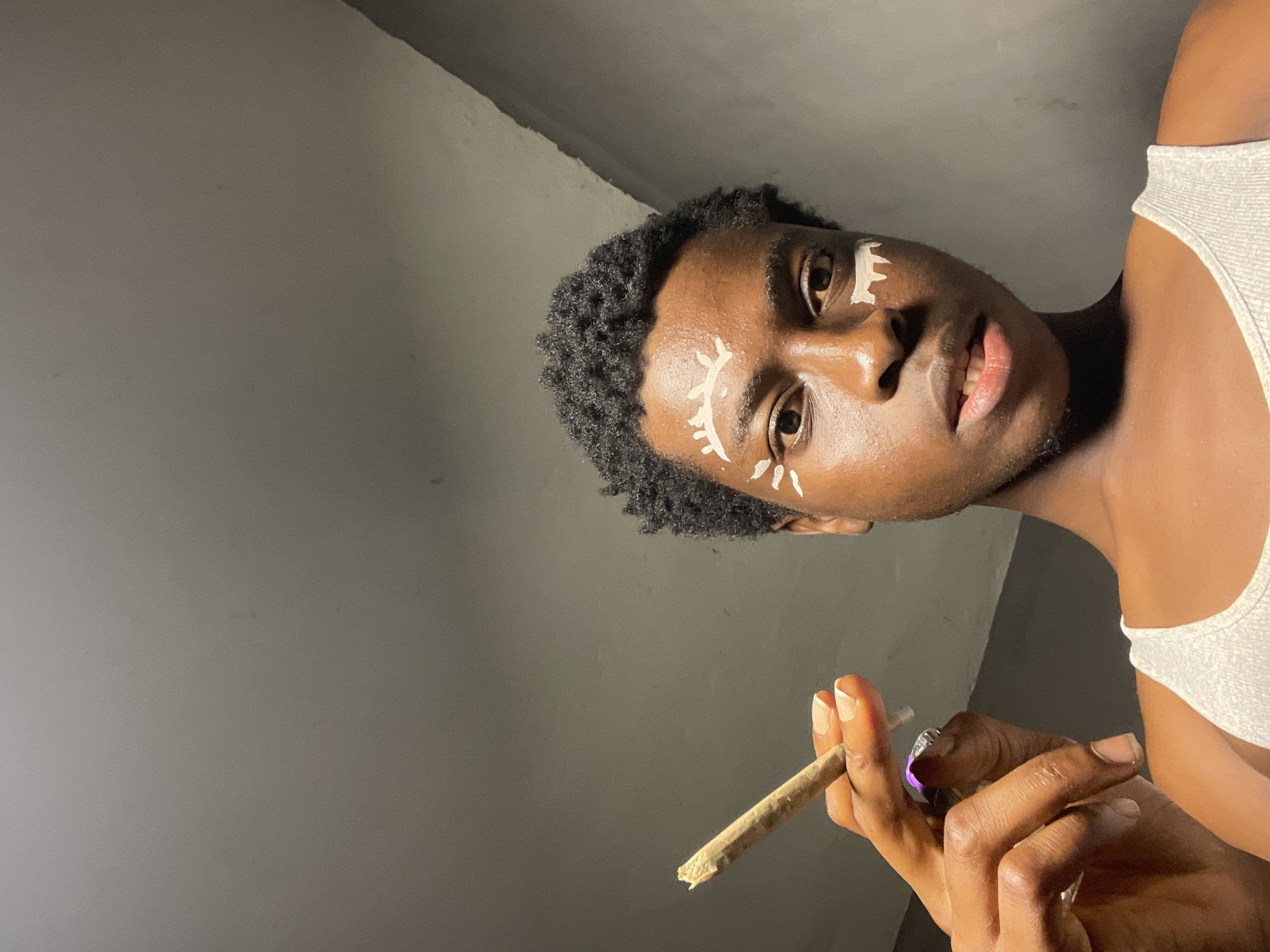 In Nkpor, Nigeria, a person with dark skin and short, curly black hair, adorned with white face paint resembling stylized eyes, holds a lit cigarette between their fingers. The cigarette is held at an upward angle, and the person is wearing a white tank top. The background consists of a neutral, slightly textured wall.