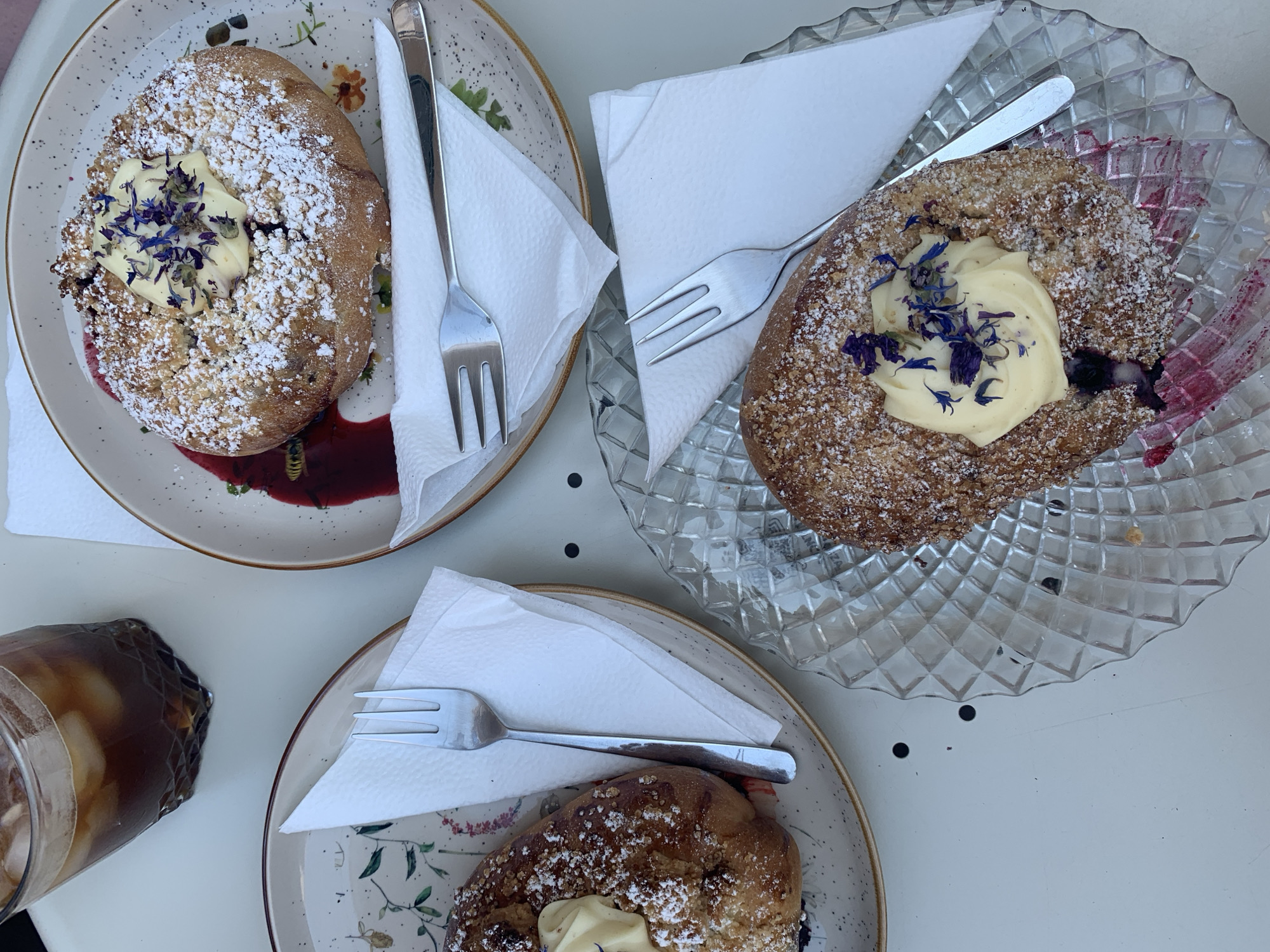 A top-down shot of three pastries with ice coffee in Poznan, Poland. The pastries are circular, topped with a dollop of yellow cream and sprinkled with powdered sugar and blue petals. One pastry is on a clear glass plate, while the other two are on ceramic plates with a floral design. Forks are placed on white napkins next to each pastry. The ice coffee is in a clear glass with ice cubes and a dark liquid. The scene is set on a white table with small black dots.