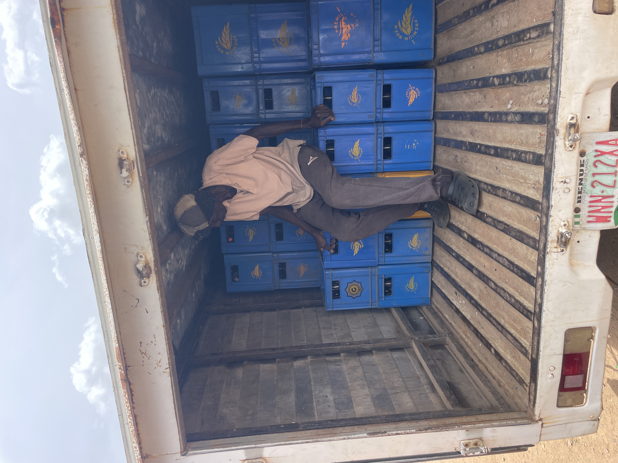 The scene shows the interior of a delivery truck, possibly in Bukuru, Nigeria. A man is lying on top of stacks of blue plastic crates labeled "PREMIUM BREW". He is wearing a light peach-colored shirt, gray pants, and black clogs. The interior of the truck is white and shows signs of wear and dirt. In the lower right corner of the image, a vehicle license plate is visible, reading "WNN-212XA" with the description "BENUE FOOD BASKET OF THE NATION".