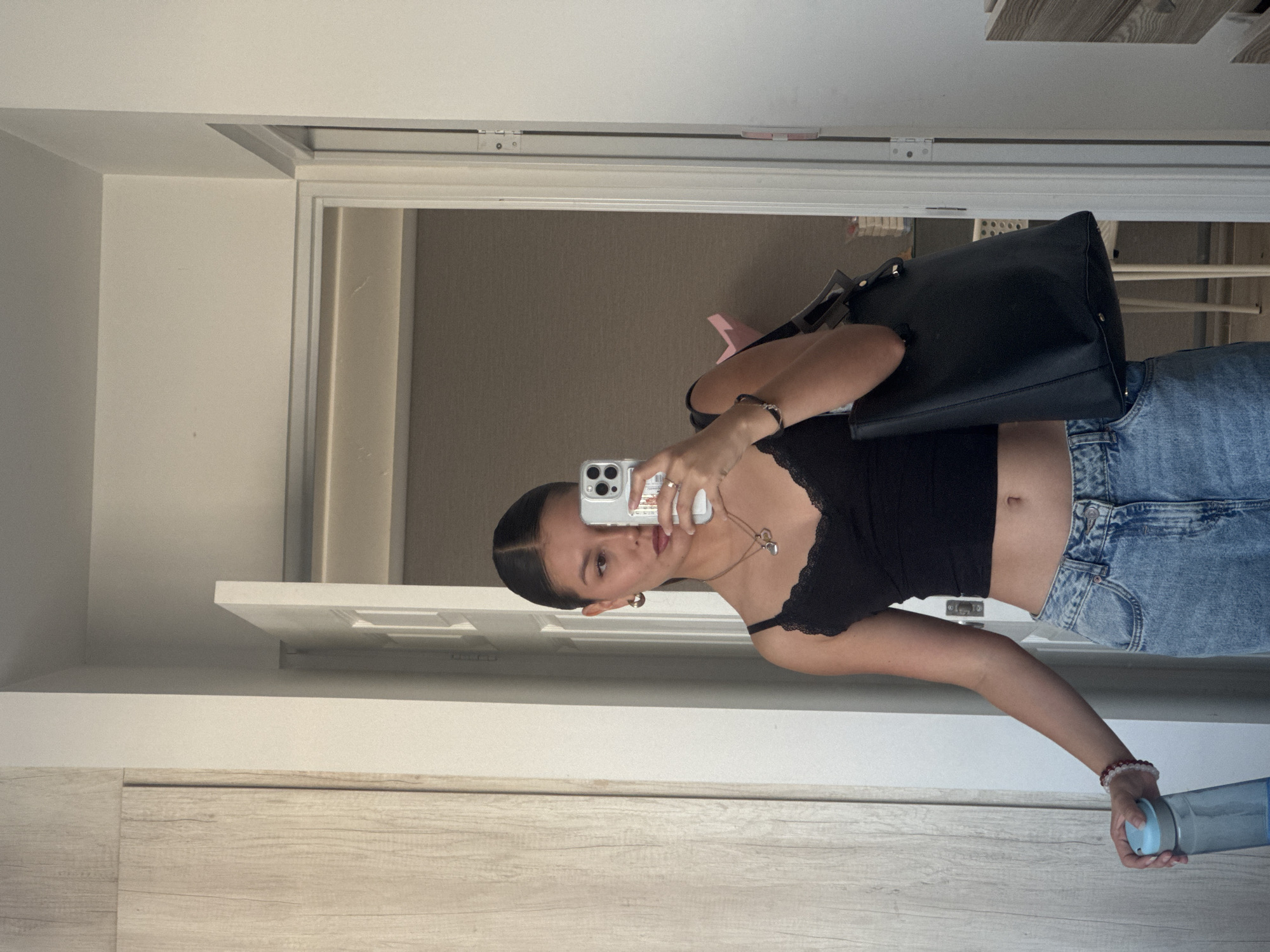 A woman stands in front of a mirror, taking a selfie with her phone. She wears a black, lace-trimmed crop top and light wash jeans. A black bag hangs from her shoulder. She holds a blue water bottle in one hand. The reflection shows an interior space with a door and beige walls. The location is in San Francisco Ocotlán (Ocotlán), Mexico.