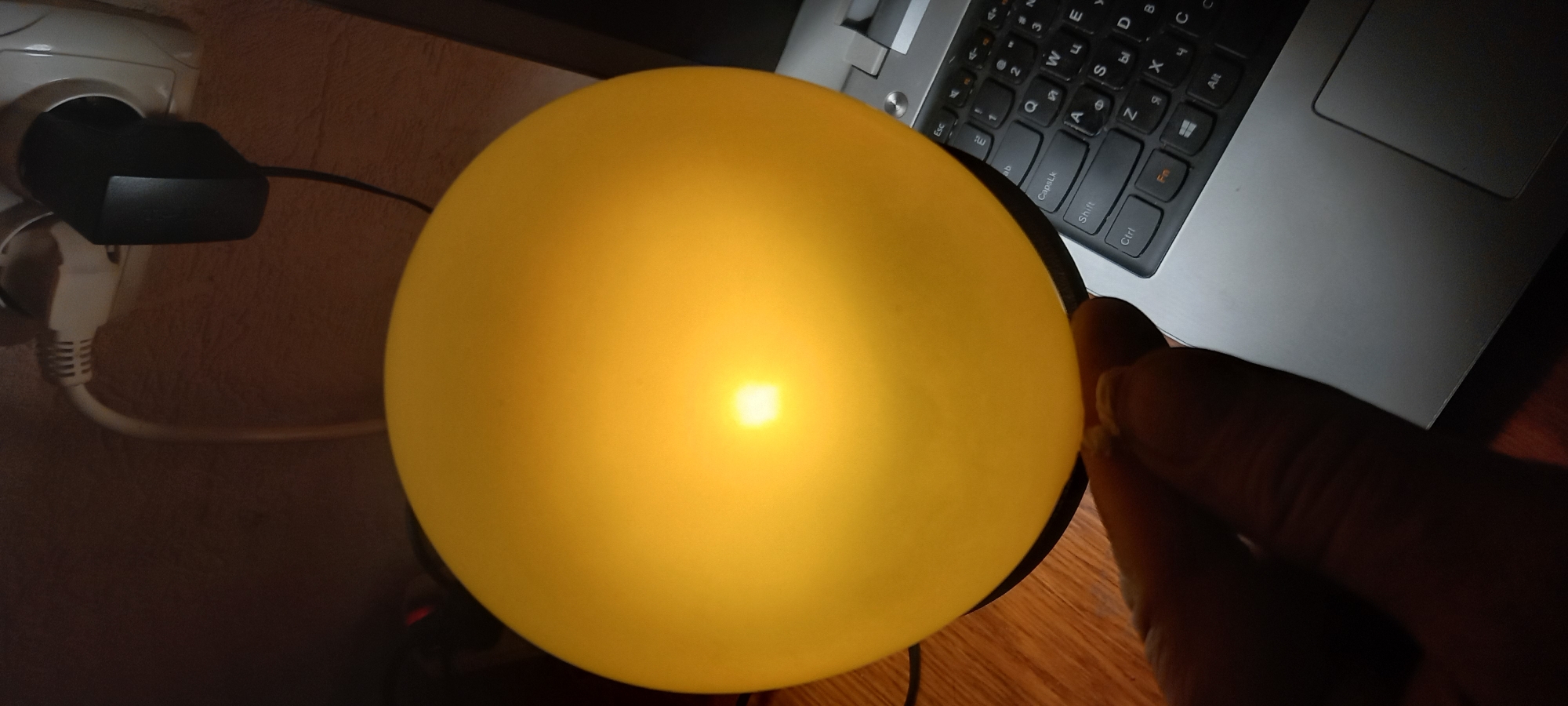 The image captures an indoor, low-light setting, likely an evening or night scene at a desk in Kryvyi Rih, Ukraine. The primary subject is a glowing, spherical yellow object, possibly a lamp or a light inside a balloon, which casts a bright, warm light. A person's hand is visible on the right, appearing to hold or adjust this light source.
In the background to the left, a white electrical outlet is visible on a light brown textured wall. A black power adapter is plugged into the top socket, with a white power cord extending downwards. To the right and behind the glowing object, a silver laptop is open, showing a portion of its keyboard. The keyboard features both Latin and Cyrillic characters, typical for Ukrainian layouts. Visible keys include "Esc", "Q", "W", "E", "CapsLk", "A", "S", "D", "Shift", "Z", "X", "C", "Ctrl", "Fn", "Alt", as well as Cyrillic characters like "Я" and others that correspond to numbers "2", "3", "4". The laptop's touchpad area is also partially visible. The objects rest on a wooden surface, likely a desk or table, which appears dark due to the low ambient light.