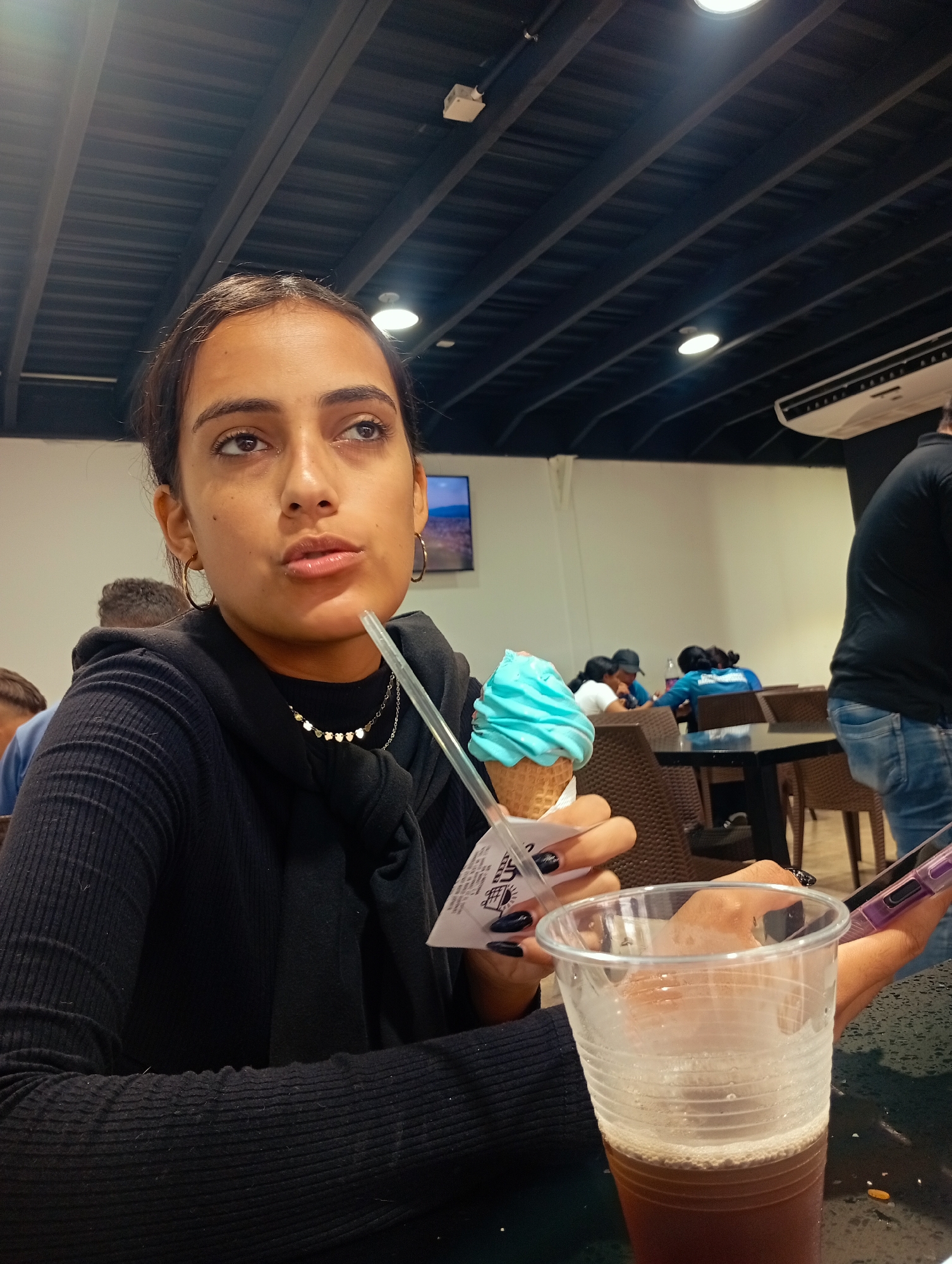The image depicts a young woman seated indoors at what appears to be a casual dining establishment or food court in Juan Griego, Venezuela. She is the main subject, positioned in the foreground, looking upwards and to her right with a thoughtful expression, her mouth slightly open. She has dark hair, dark eyes, and a medium skin tone, and is wearing a black long-sleeved ribbed top. A black scarf or hood is draped around her neck, complemented by gold hoop earrings and a delicate heart-shaped necklace. Her fingernails are painted a dark color.
In her right hand, she holds a light blue ice cream cone, which appears to be a soft-serve type, with a clear plastic spoon/straw inserted. On the table in front of her is a clear plastic cup filled with a dark brown beverage, possibly iced tea or soda, topped with a layer of foam. A mobile phone with a purple case is partially visible in her left hand.
The background reveals a bustling environment with other people seated at dark tables and chairs. A person in a black t-shirt and blue jeans is partially visible on the far right. The ceiling is dark with visible beams or panels and features several recessed lights. A white air conditioning unit is mounted high on the wall to the right, and a screen or television is visible in the far distance. The scene is illuminated by artificial lighting. Illegible text is visible on the paper wrapper around the ice cream cone.