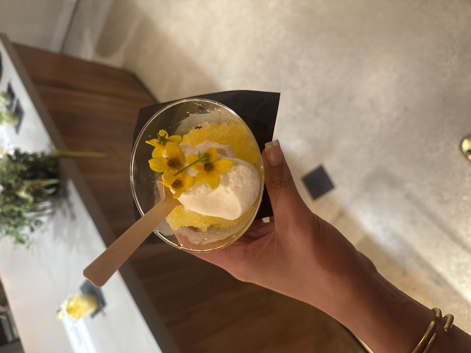 An indoor close-up photograph taken in Los Angeles, United States of America, depicts a person's right hand holding a dessert. The hand, with light to medium skin tone and short, manicured nails, holds a transparent plastic cup containing a bright yellow granular substance, likely a granita or sorbet, topped with a scoop of white cream or ice cream. Several small yellow flowers with dark centers garnish the cream, and a light-colored wooden spoon is inserted into the dessert. A gold-colored, open-cuff bracelet is worn on the subject's wrist. The dessert cup rests on a dark, possibly black, napkin. The background features a light grey countertop with a wooden base, exhibiting a slanted panel design. Out-of-focus green foliage and additional small yellow items, similar to the held dessert, are visible on the countertop. The floor is a light-colored, possibly concrete or tiled, surface with a dark square inlay visible in the lower portion of the frame.