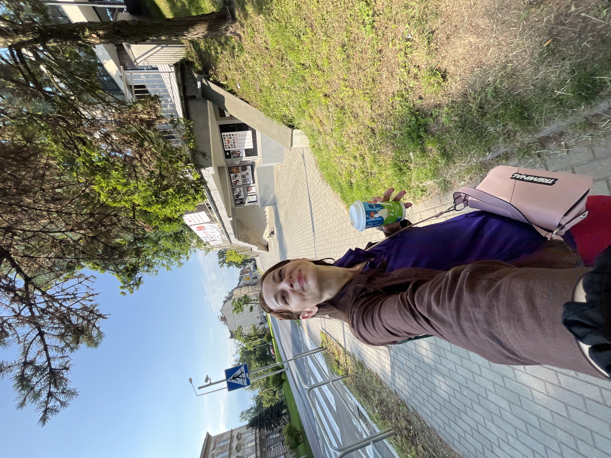 A young woman is walking on a paved sidewalk next to a street in a city. She is holding a disposable coffee cup in her right hand and a pink purse is slung over her left shoulder. She is looking up and to her right with a smile. She is wearing a purple shirt and a brown jacket. Behind her, across the street, there is a building with several floors, and to her right, there is a grassy slope with trees. The sky is blue with some clouds.