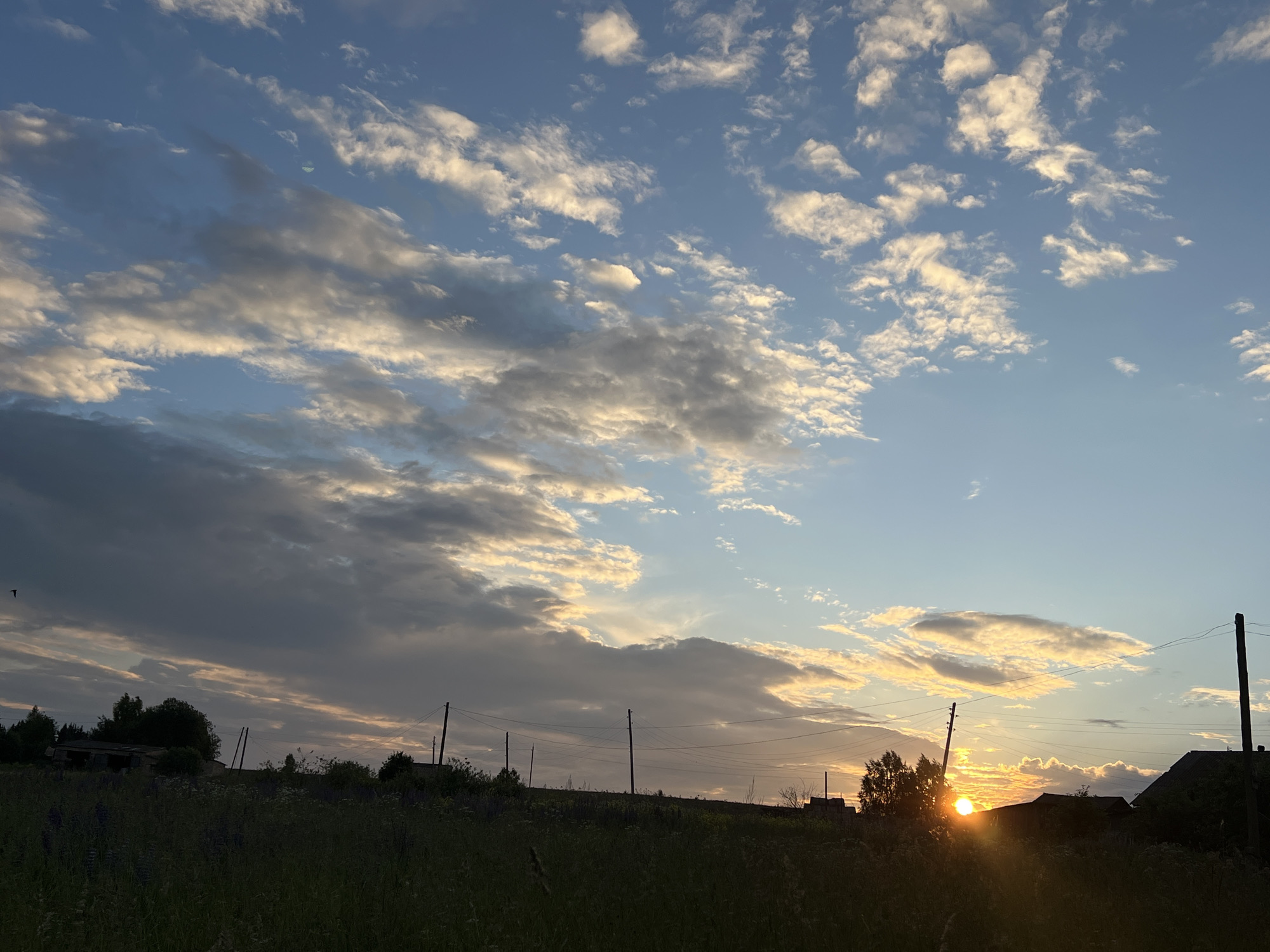 This image captures a sunset in Panteleevka, Russia. The sky is filled with a mix of light and dark clouds, with the sun low on the horizon, casting a warm glow. In the foreground, a dark silhouette of a grassy field with some plants and trees is visible. On the right side of the image, a portion of a house and some utility poles with wires are present. The overall scene depicts a rural landscape during the golden hour.