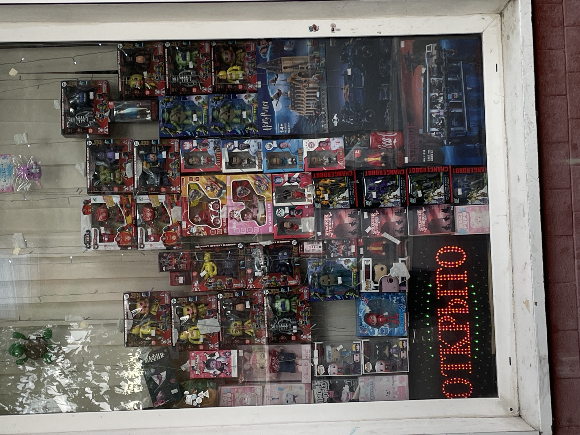 This media file is a photograph of a storefront display filled with various toys and collectibles. The display is behind a glass window, which reflects some of the surroundings, including foliage and possibly sunlight. The arrangement of items suggests a toy store or a similar retail establishment.
The subjects are primarily objects, specifically packaged toys. There are many different types of toys visible, including action figures, dolls, building sets (such as a "Harry Potter" castle and a "Sonic the Hedgehog" themed item), and other novelty items. Some of the toys appear to be from popular franchises like "Stranger Things." The sheer volume and variety of toys create a sense of abundance and excitement, typical of a children's store.
There are no people actively engaged in any activity within the frame of the image, but the display itself is the focal point. The setting is clearly an indoor display within a store, viewed from the outside.
Notable details include a bright, illuminated sign in the lower right corner that reads "ОТКРЫТО" (OTKRYTO), which means "OPEN" in Russian, confirming this is a retail space. The text on the toy packaging is also visible, with titles like "Harry Potter," "Stranger Things," "Sonic the Hedgehog," and other unidentifiable product names and brandings in various languages, including Russian. The time of day appears to be daytime, given the lighting and reflections on the glass. The weather is not directly discernible, but the reflections suggest it is likely not raining heavily. The presence of Russian text on the "OPEN" sign indicates the location is in Russia, likely Moscow as per the context. The overall impression is a vibrant and busy retail display designed to attract customers.